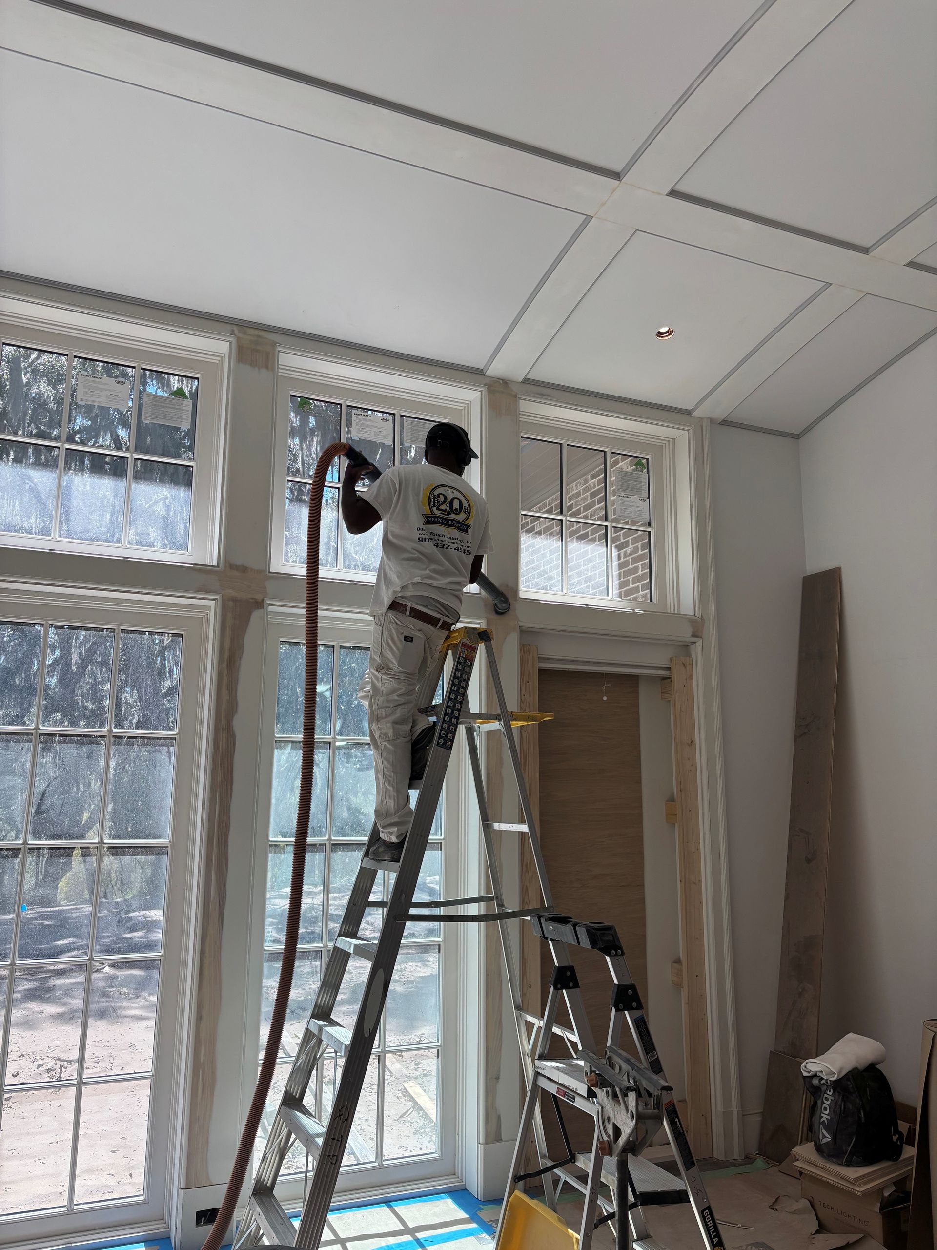 Person on ladder spraying paint near large windows. Interior setting with white walls and ceiling.