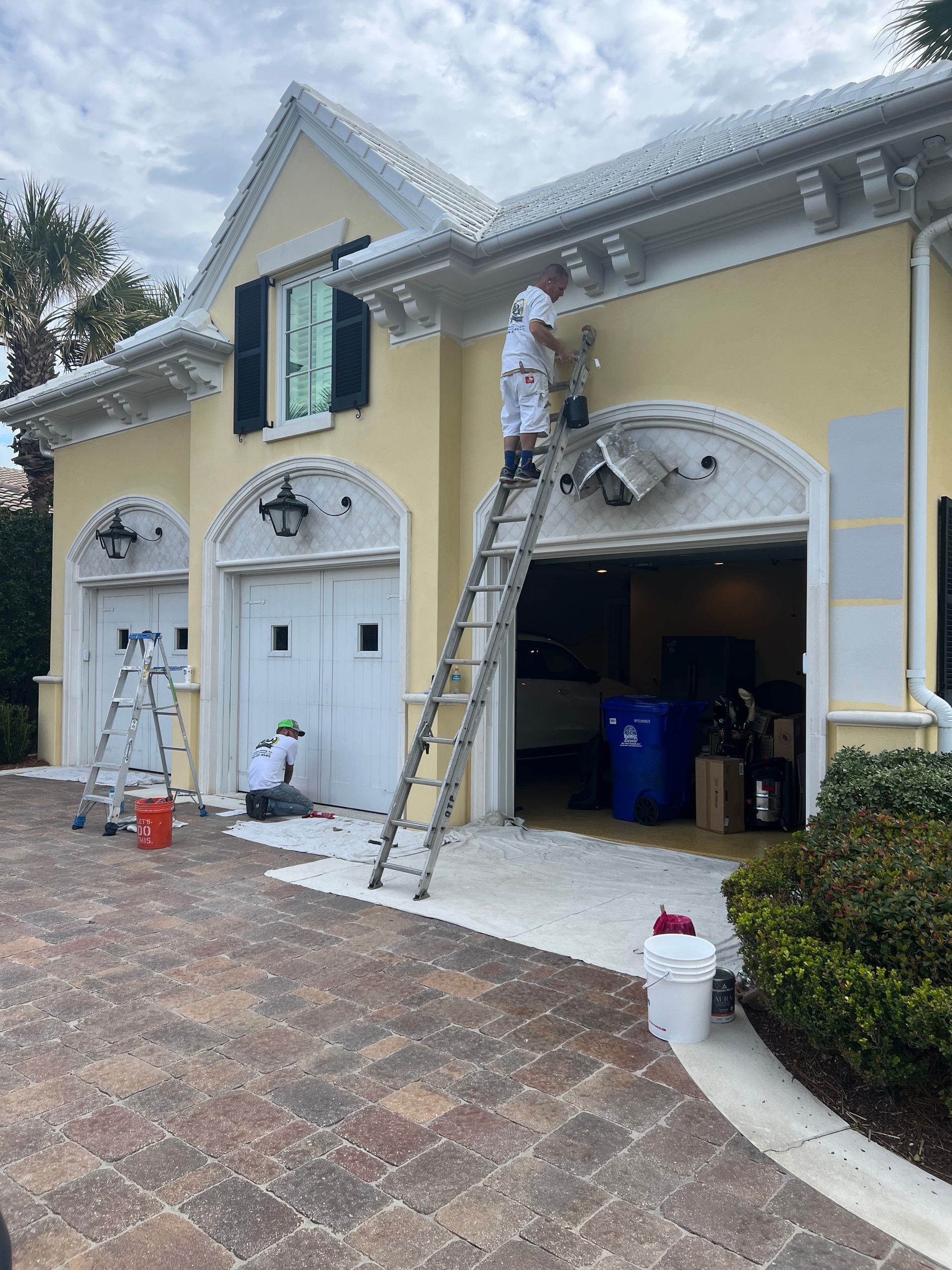 Workers on ladders painting a yellow house exterior with white trim and garages.