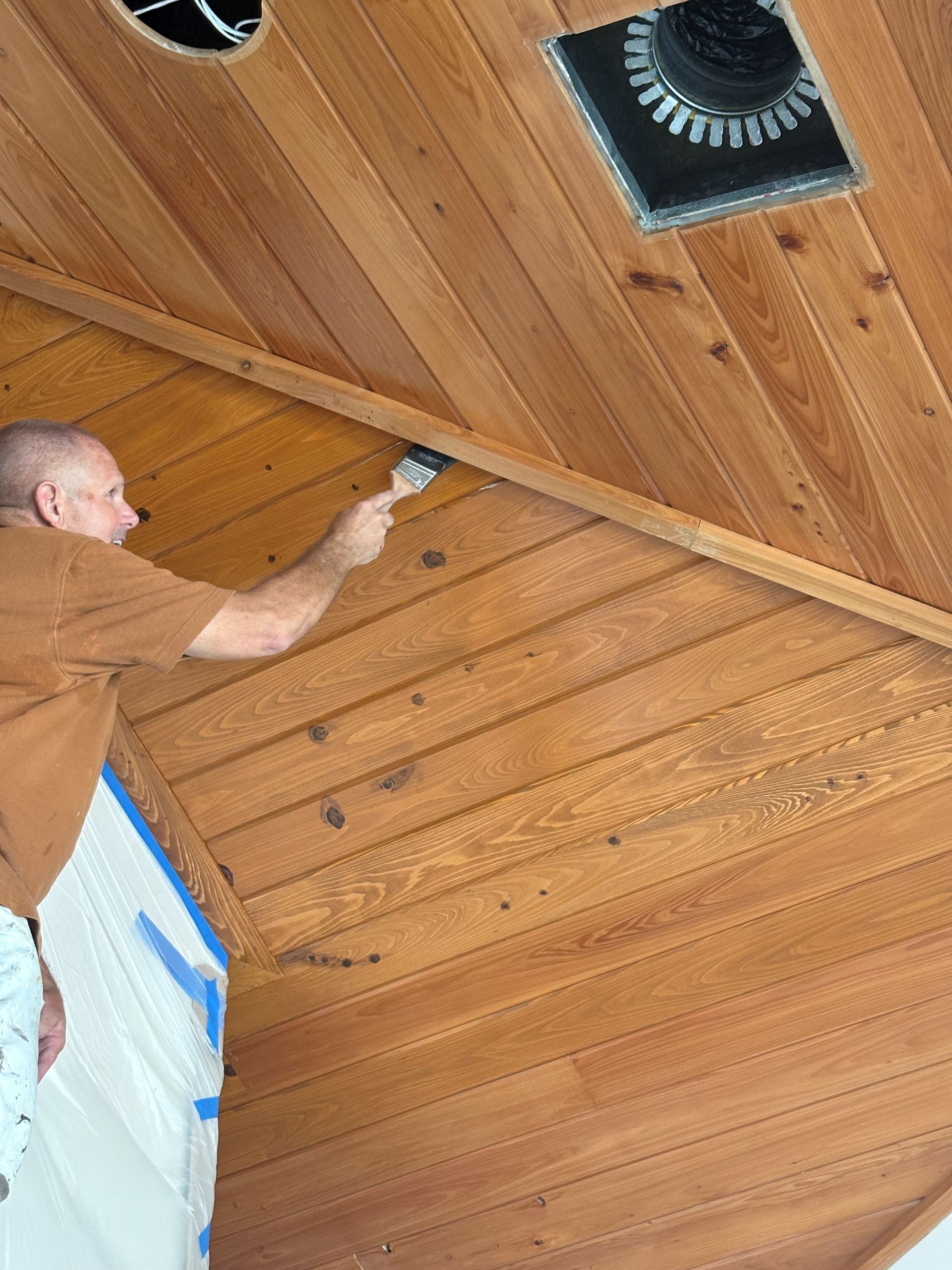 Man painting a wooden ceiling with a brush. The wood is warm brown.
