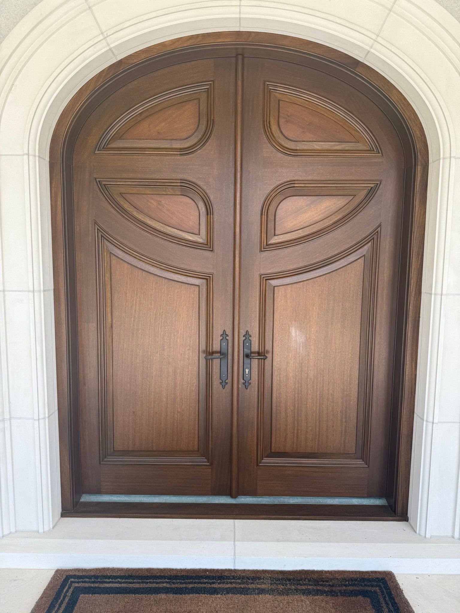 Double wooden arched front doors with decorative carvings and black hardware, set in a white stone frame.