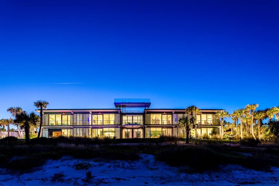 Modern beachfront house at dusk, illuminated interior, dark blue sky.