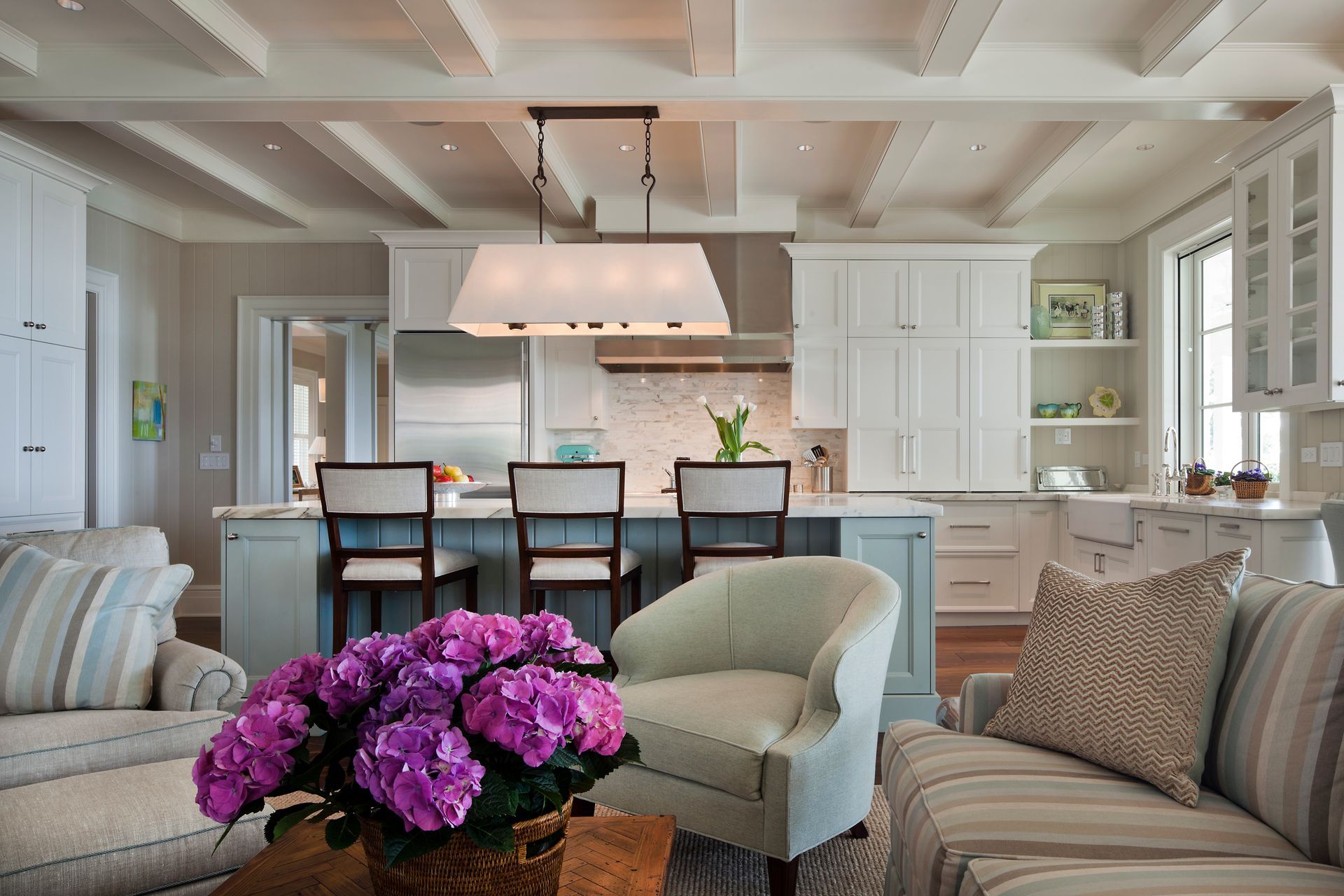 Coastal-style kitchen with an island, bar stools, and a seating area with a blue armchair and a sofa.