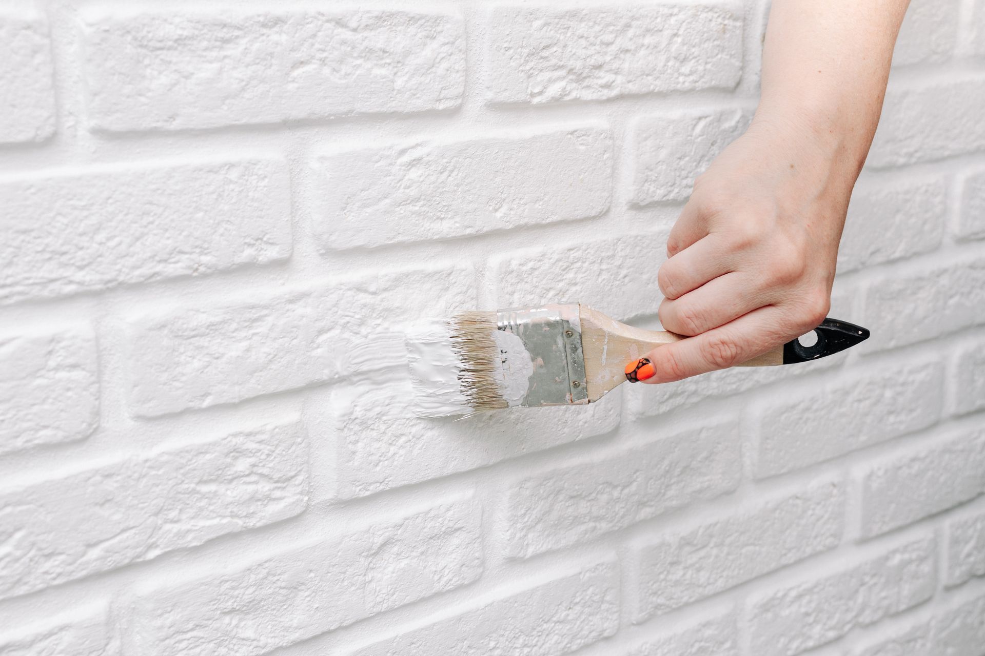 Hand using a paintbrush to apply white paint to a brick wall