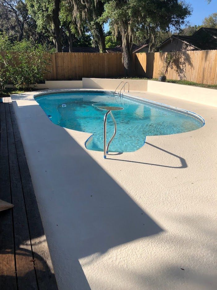 Pool with light-colored deck in a backyard, surrounded by a wooden fence and trees.