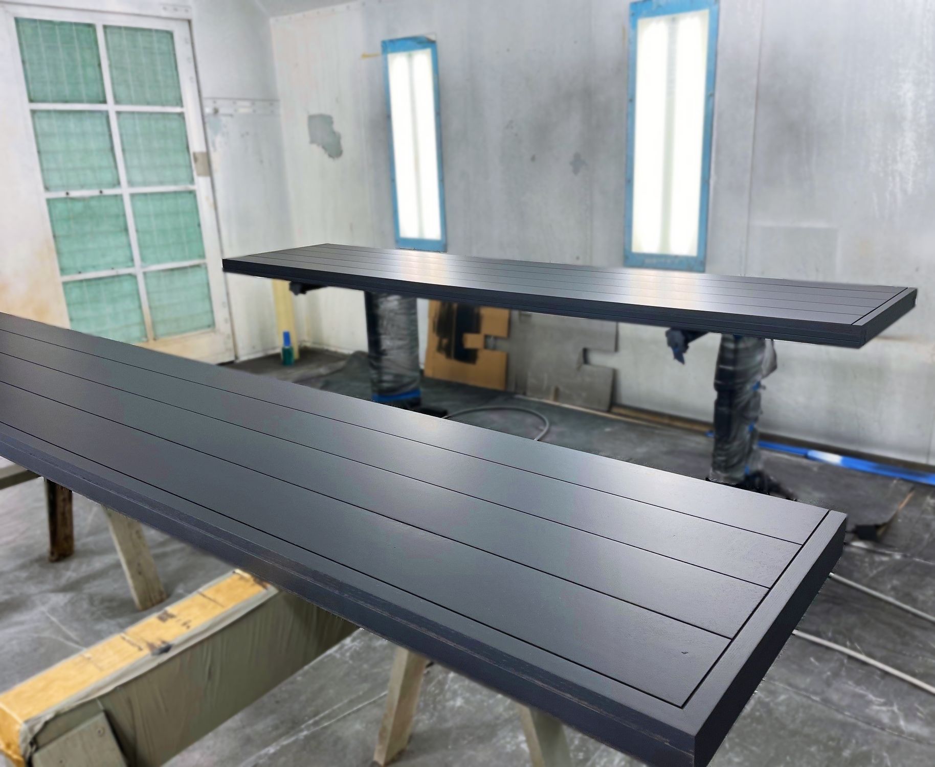 Two large, dark gray tables in a spray painting booth, one on sawhorses, the other with covered legs.