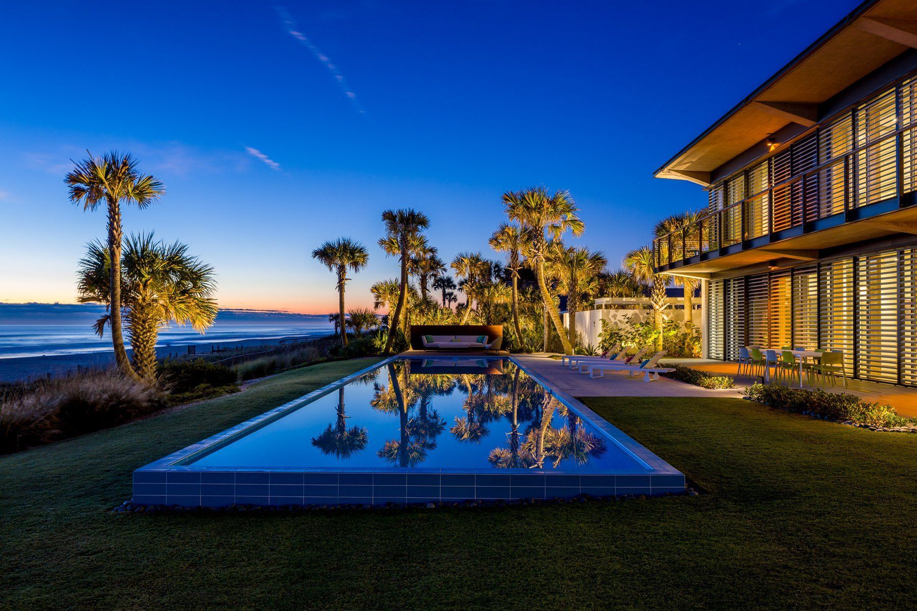 Night view of a luxurious house with a long pool, palm trees, and the ocean in the background.