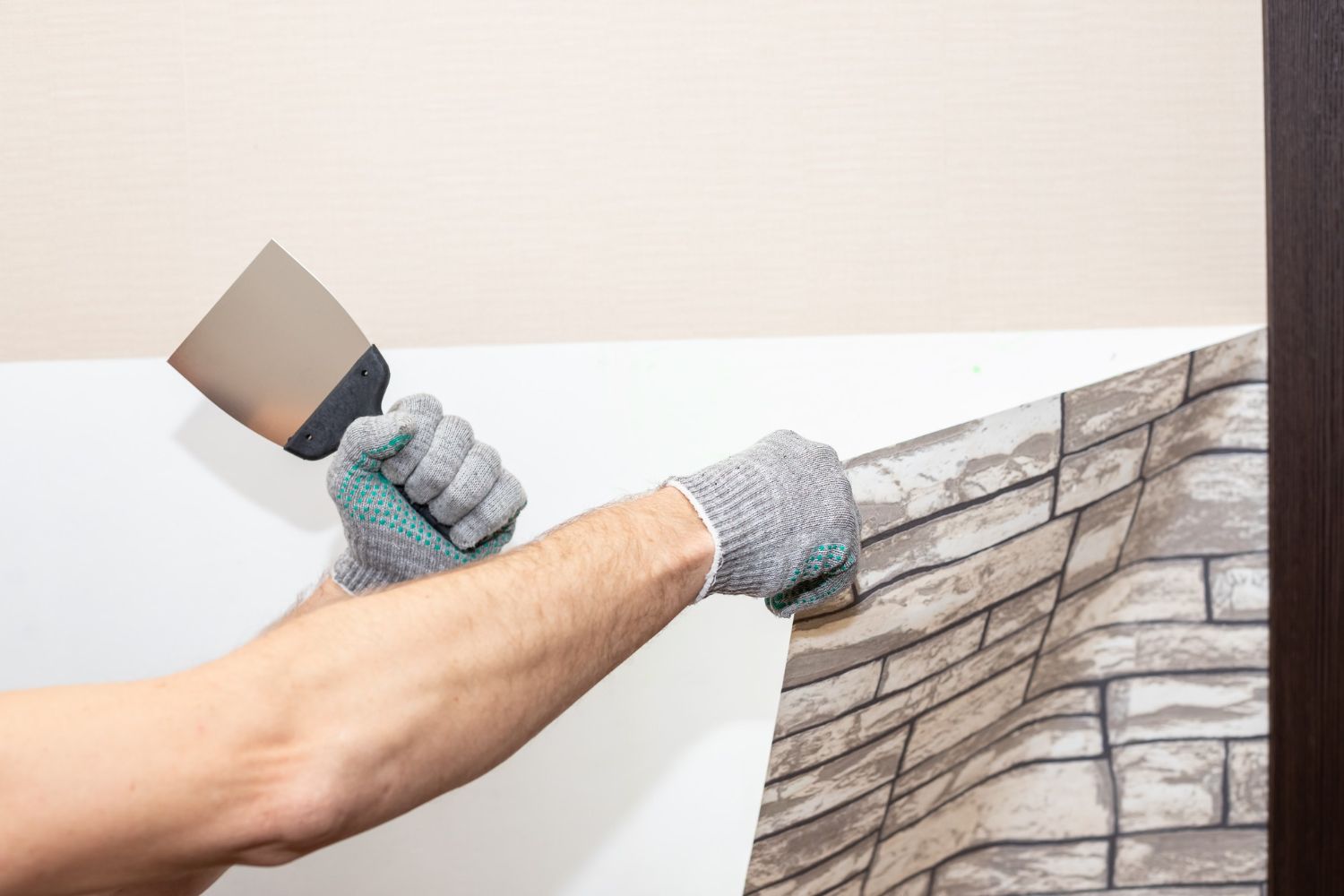 Person removing brick-patterned wallpaper from a white wall using a putty knife, indoors.