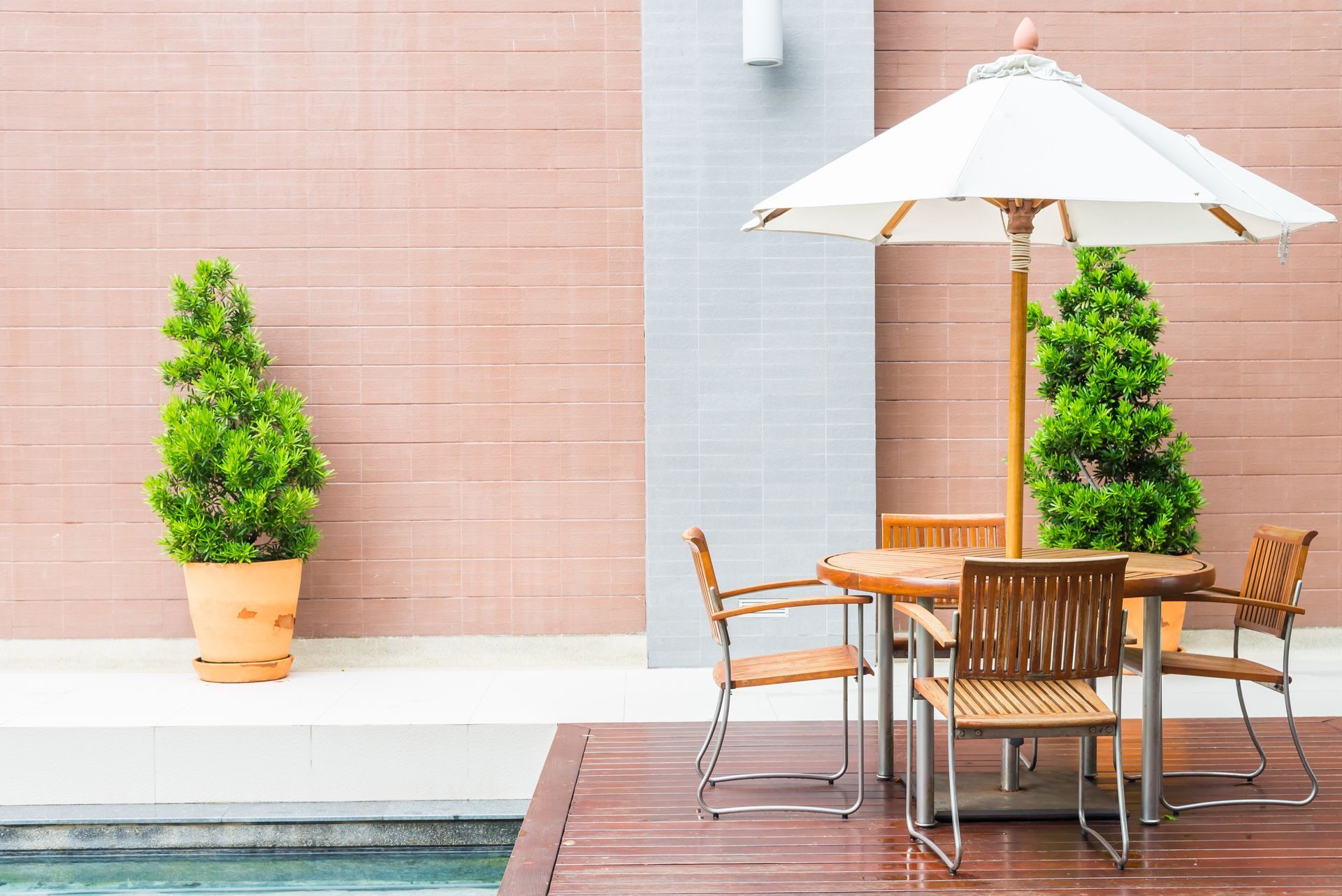 Modern patio with wooden table, chairs, umbrella, and potted green plants beside a pool
