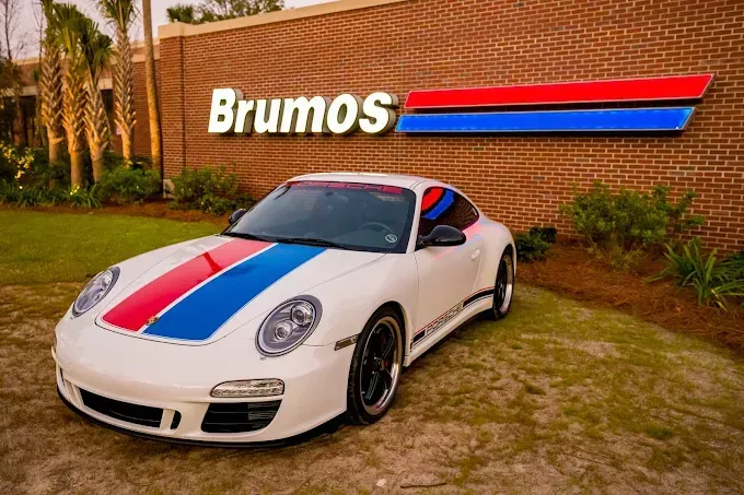 White Porsche with red, blue, and white racing stripes parked outside Brumos dealership with brick facade.