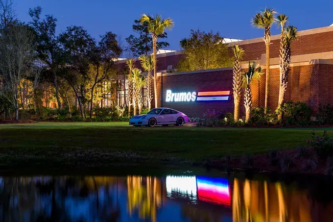 A white Porsche sports car parked in front of a Brumos sign at dusk, with its reflection in a pond.
