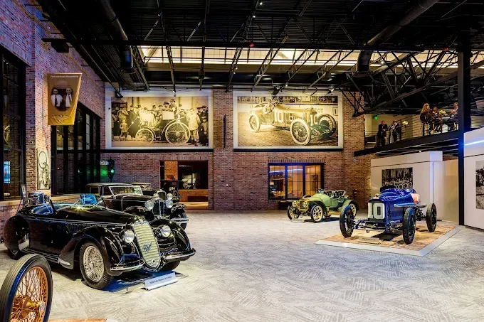 Classic car museum interior with vintage vehicles on display; brick walls, large photos, and overhead pipes.
