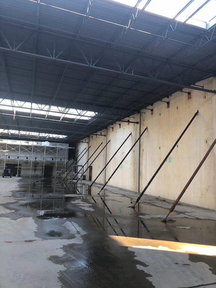 Interior of an empty warehouse with a high ceiling. Walls are supported by diagonal metal braces. Wet concrete floor.