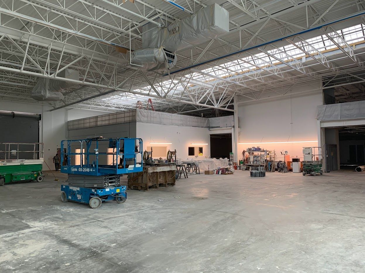 Construction site interior. White walls, metal rafters. Blue scissor lift, workers, equipment.