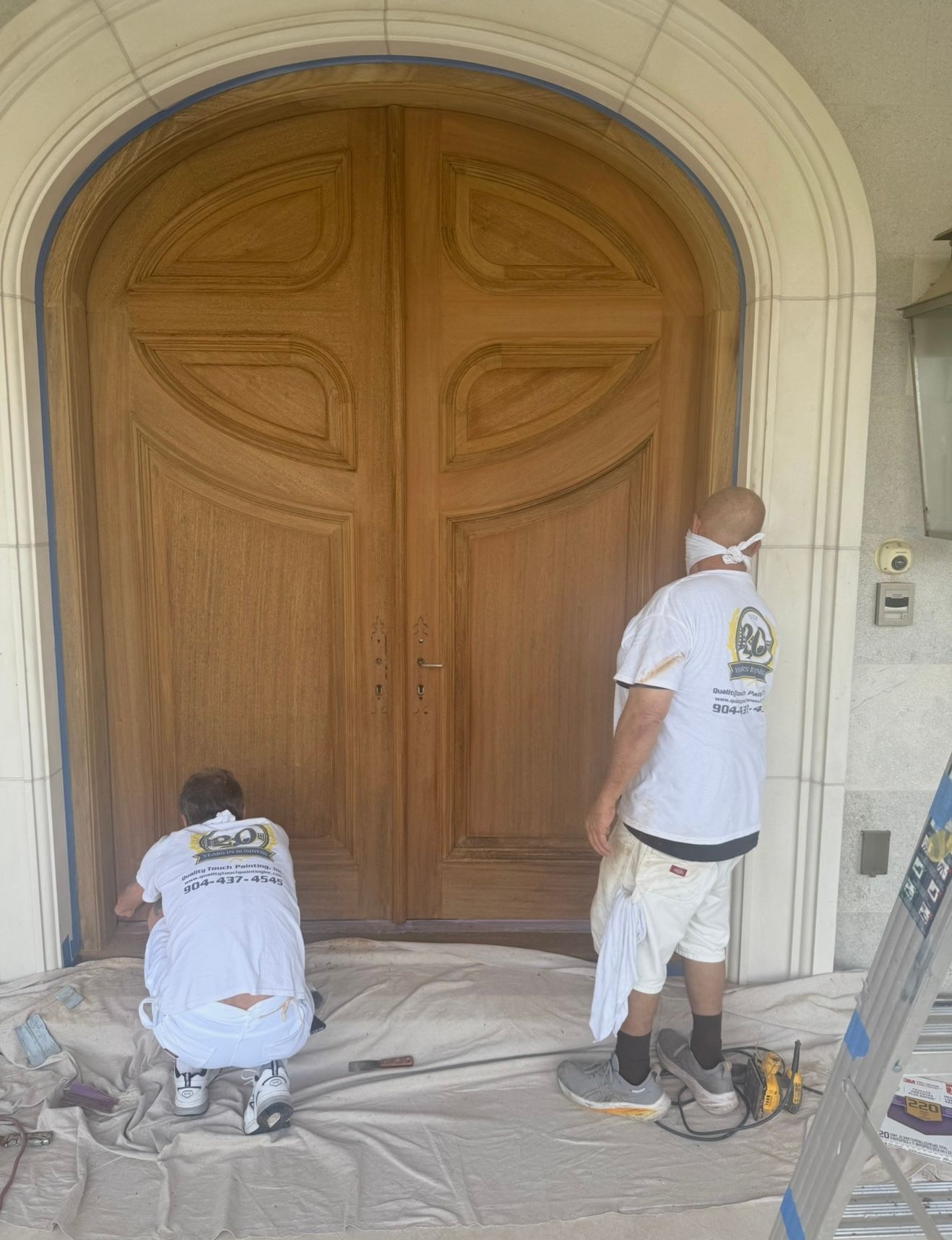 Two painters applying finish to a large wooden double door under a stone archway.