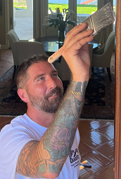Man with arm tattoo painting a wooden door frame, smiling. Inside a home.