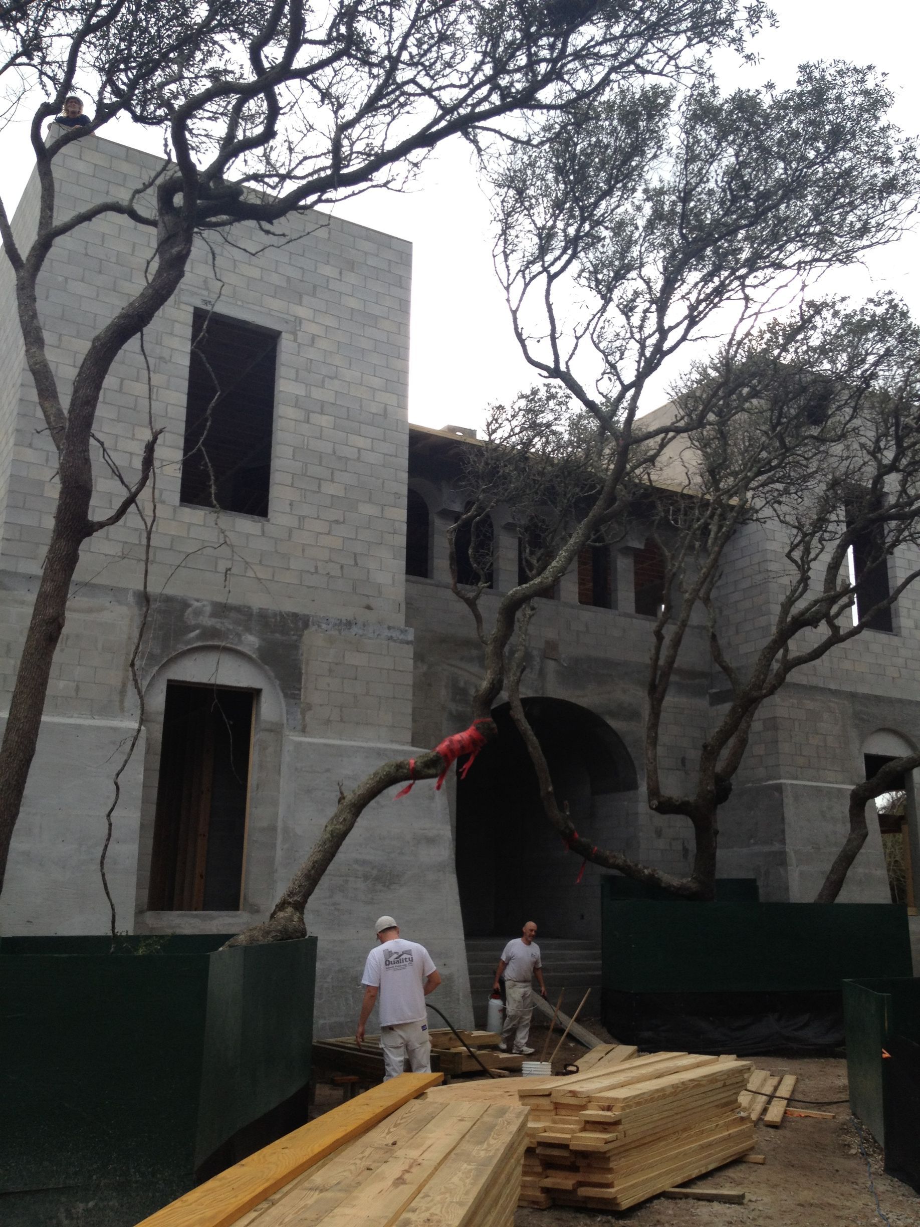 Building under construction, trees, two workers, lumber, overcast sky.