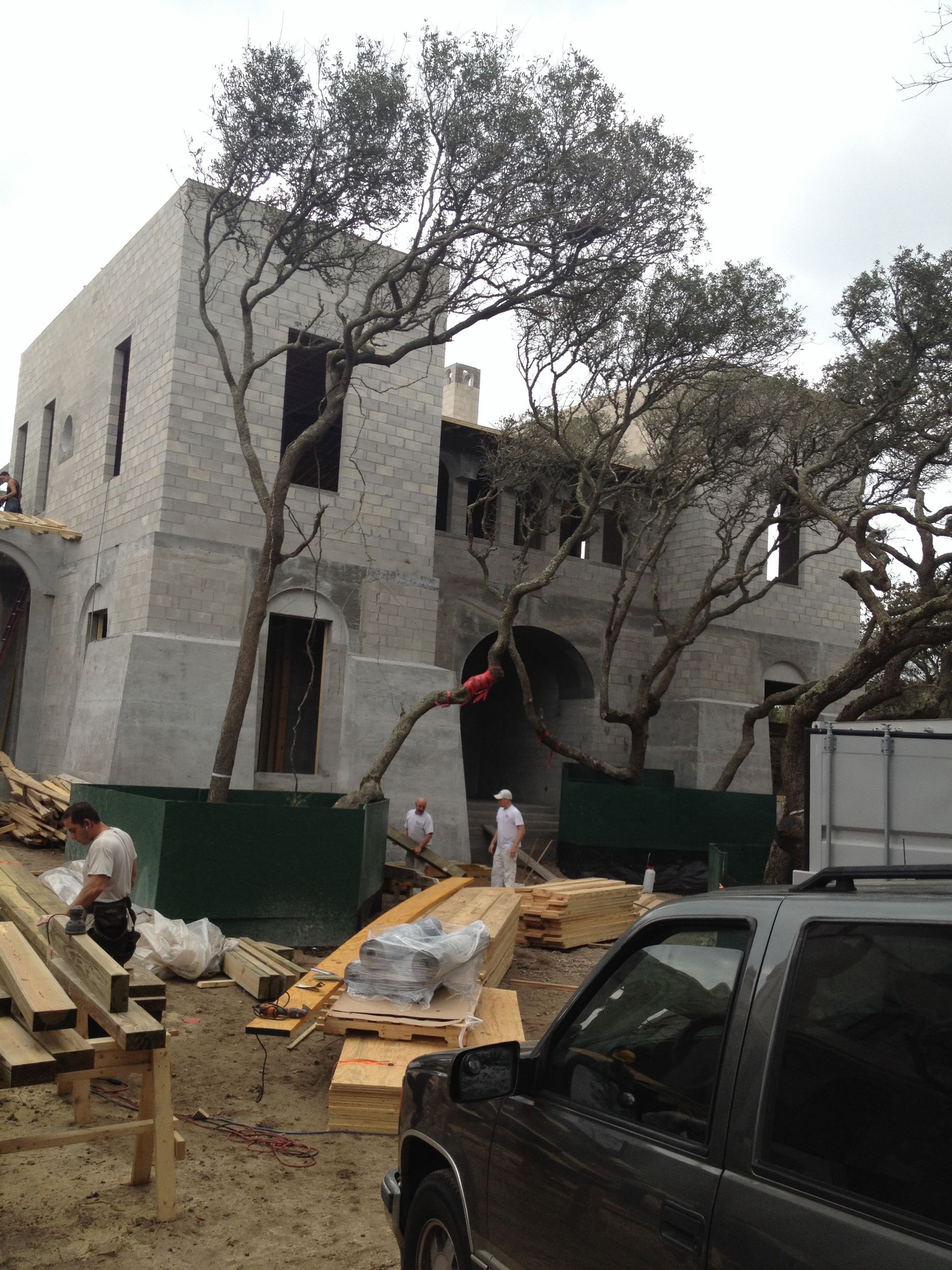 Exterior of a light-colored building under construction, trees growing through it. Workers, lumber, and truck present.