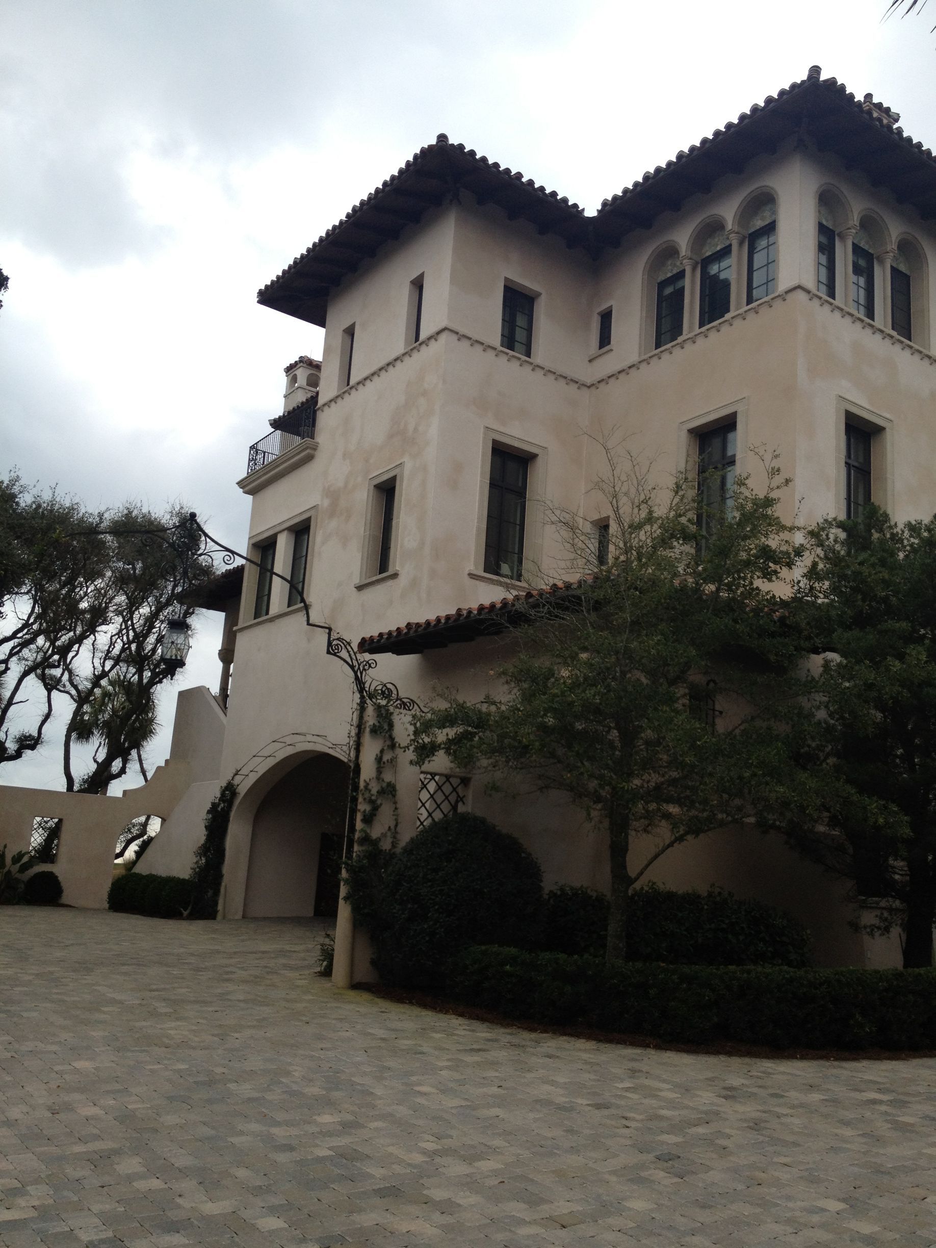 Tan stucco building with dark roof tiles, arched doorway, and small windows; cloudy sky.