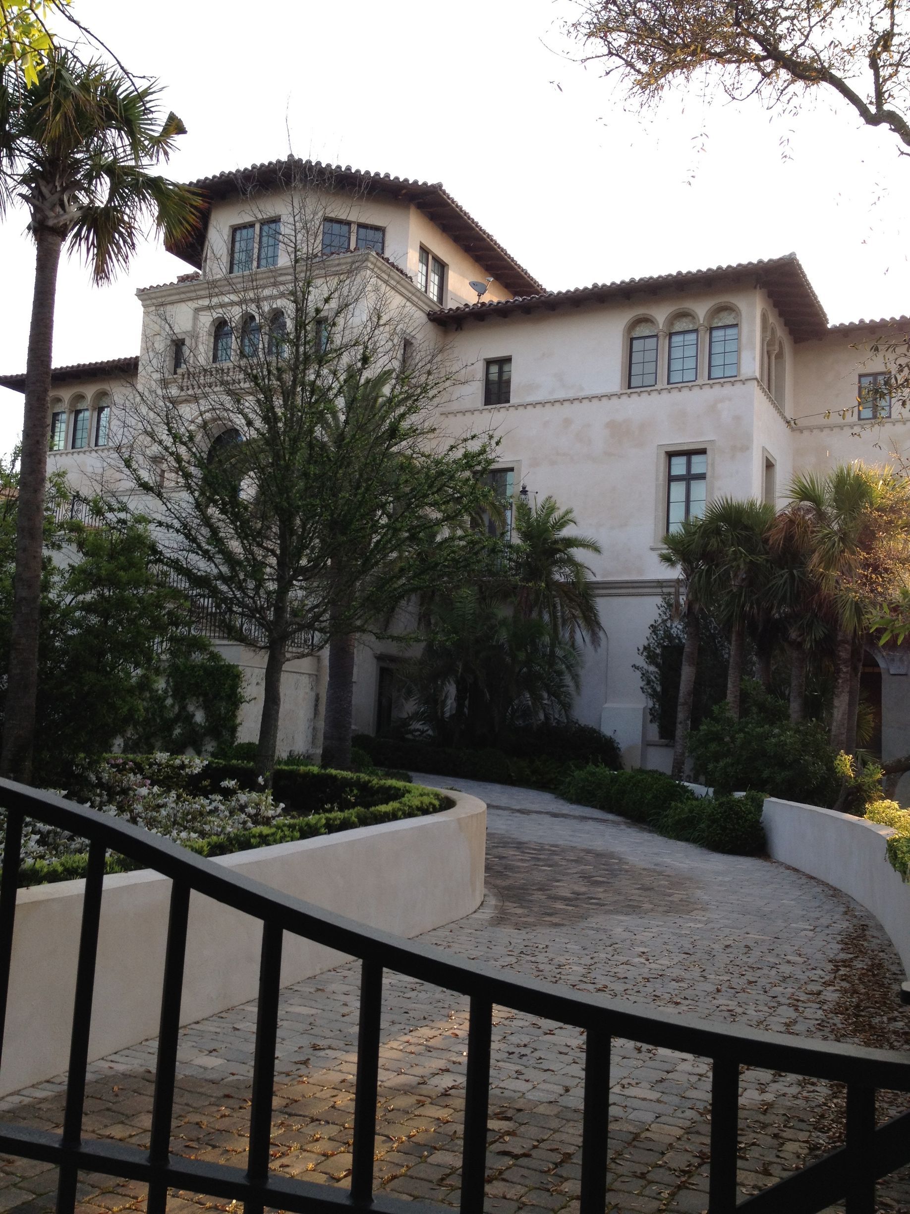 White stucco mansion with arched windows, curved driveway, and black wrought iron fence.