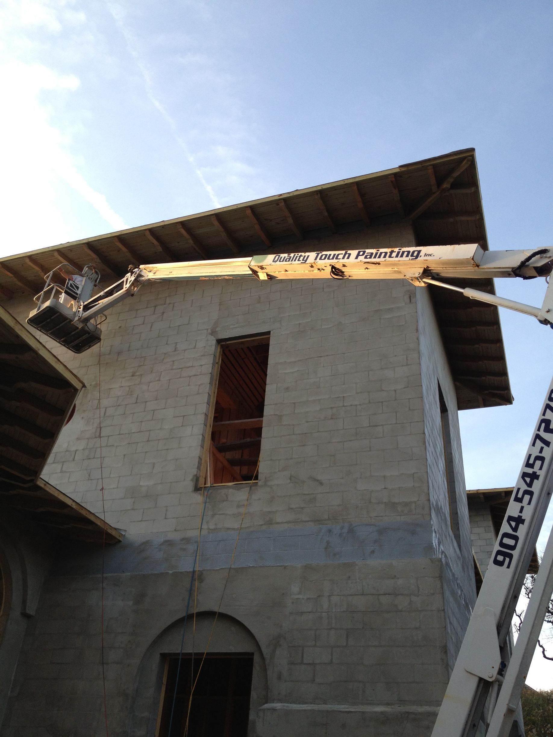 Construction worker on a lift repairing a brick building with open window frames and a wood roof, under a blue sky.