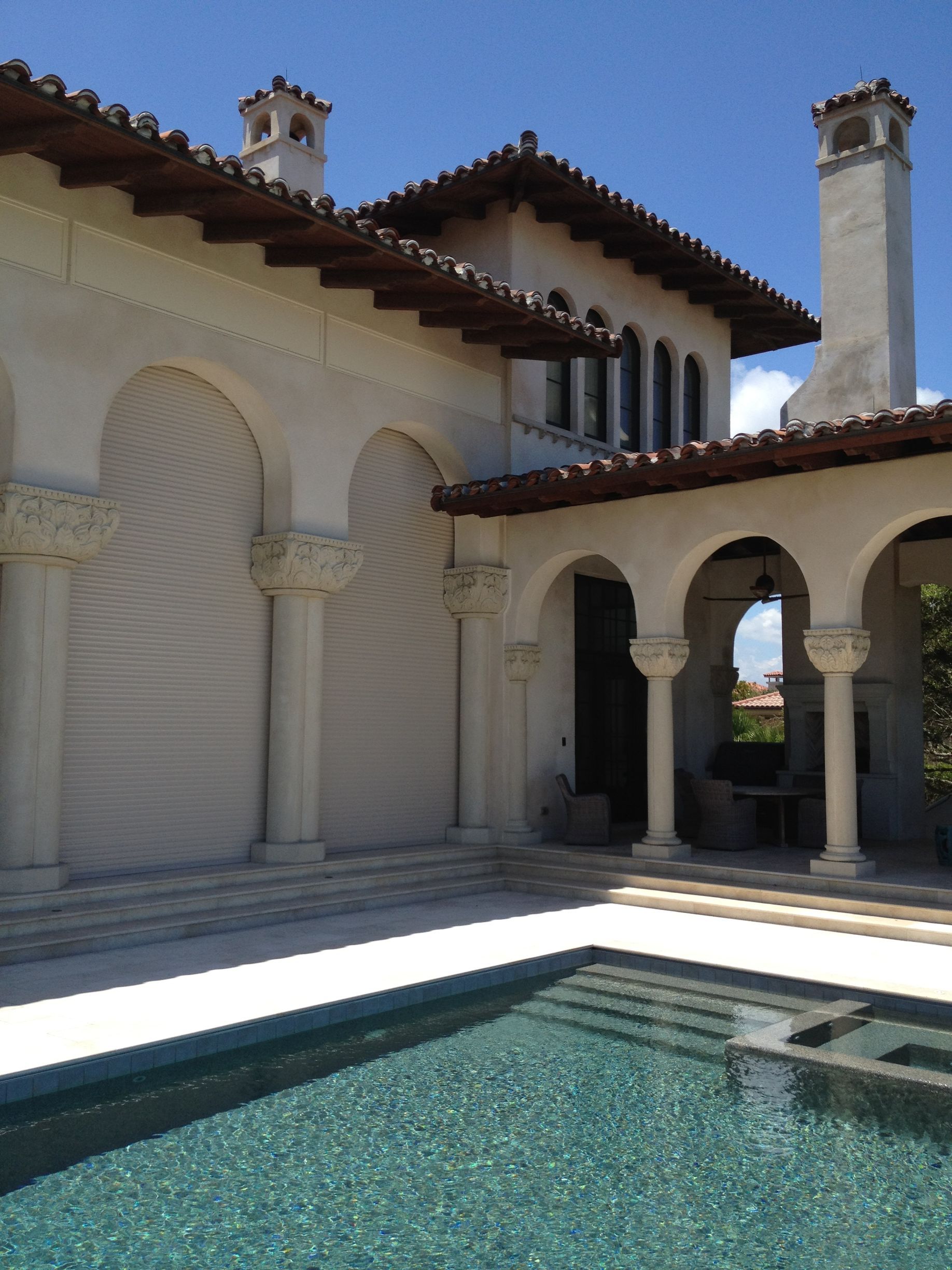 Poolside view of a luxury house with arched colonnade, light stucco walls, and blue pool under a bright sky.