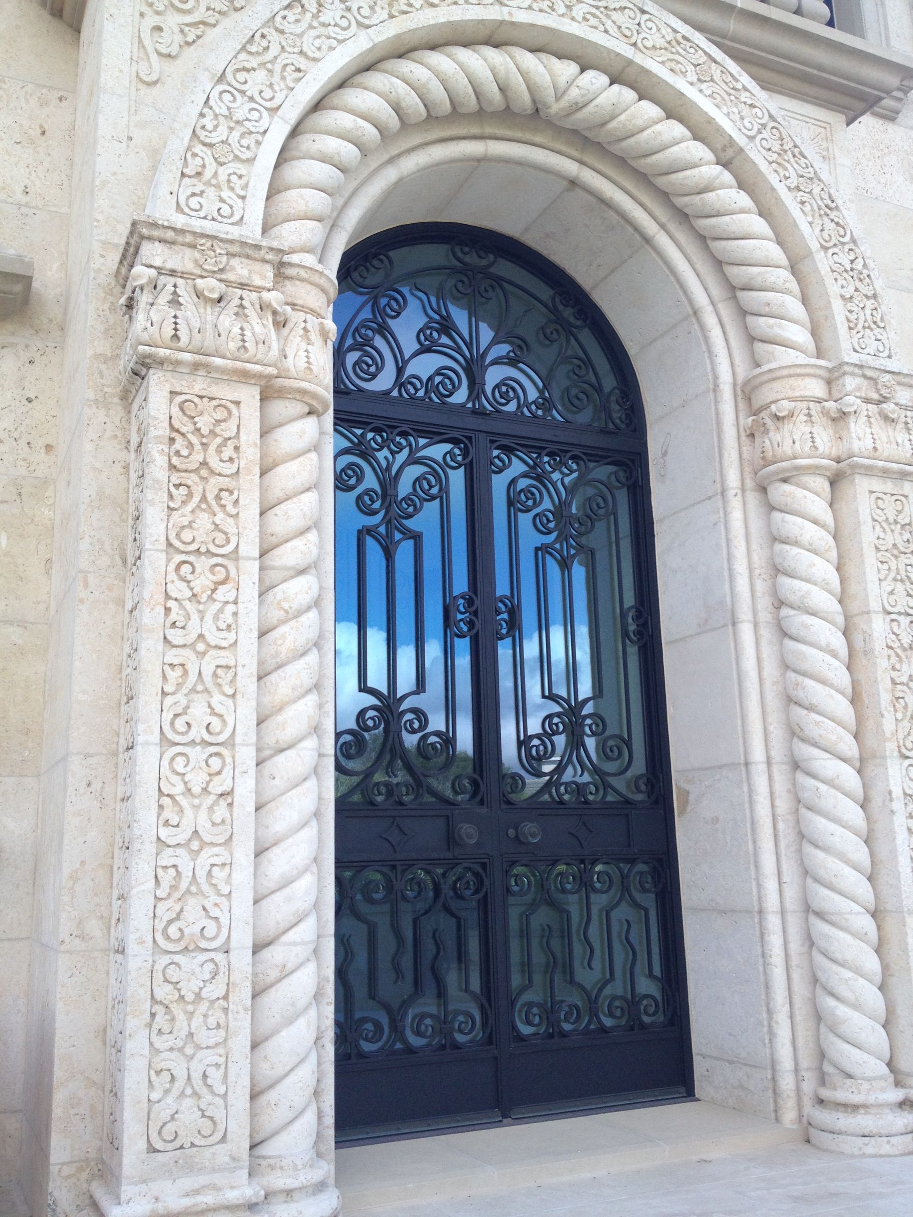 Ornate black iron doors framed by carved stone arch and pillars, against a bright sky.