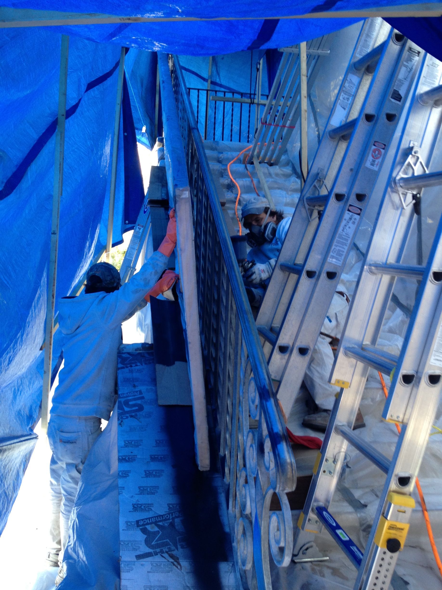Workers install railing on stairwell, inside blue tarp enclosure, using ladders.