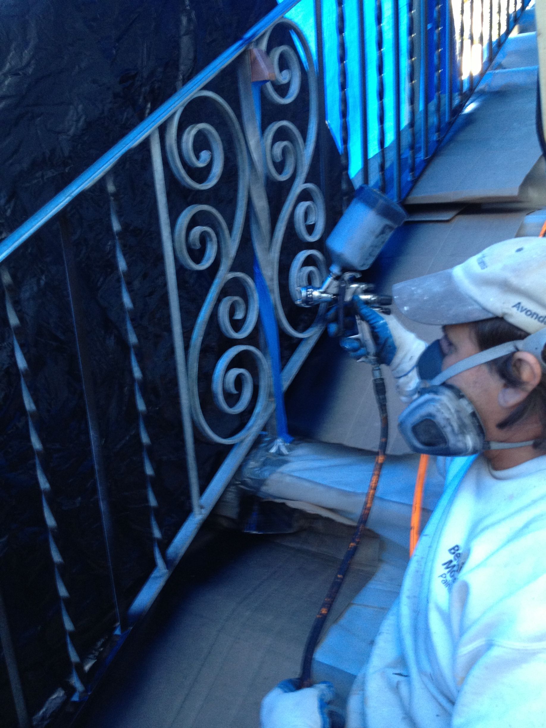 Person spray painting a wrought iron railing blue, wearing a respirator, outdoors.