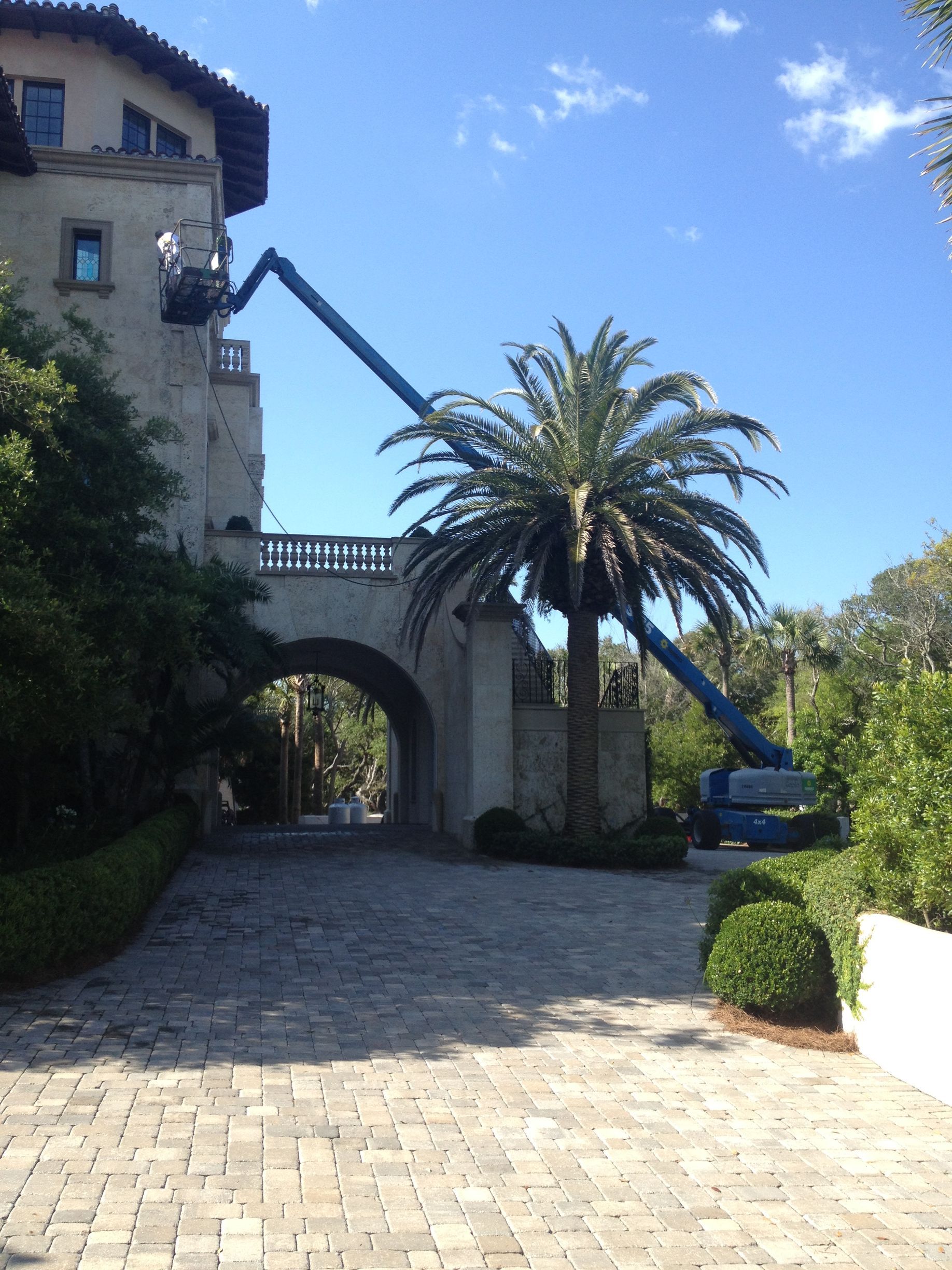 Construction crew in lift working on the facade of a large, stone building near a palm tree.