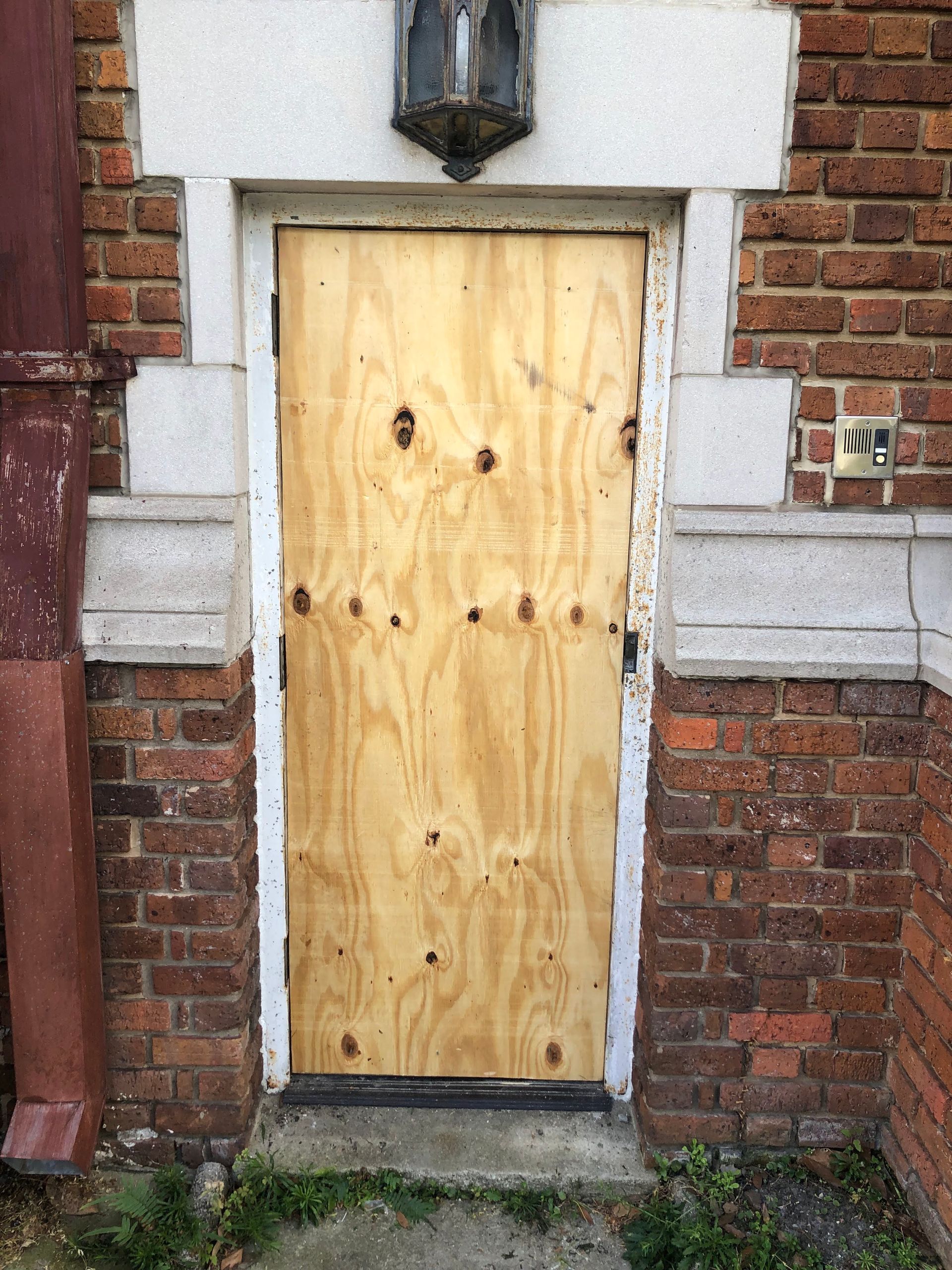 Boarded-up wooden door set in a red brick building with stone accents. Door has visible screw holes.