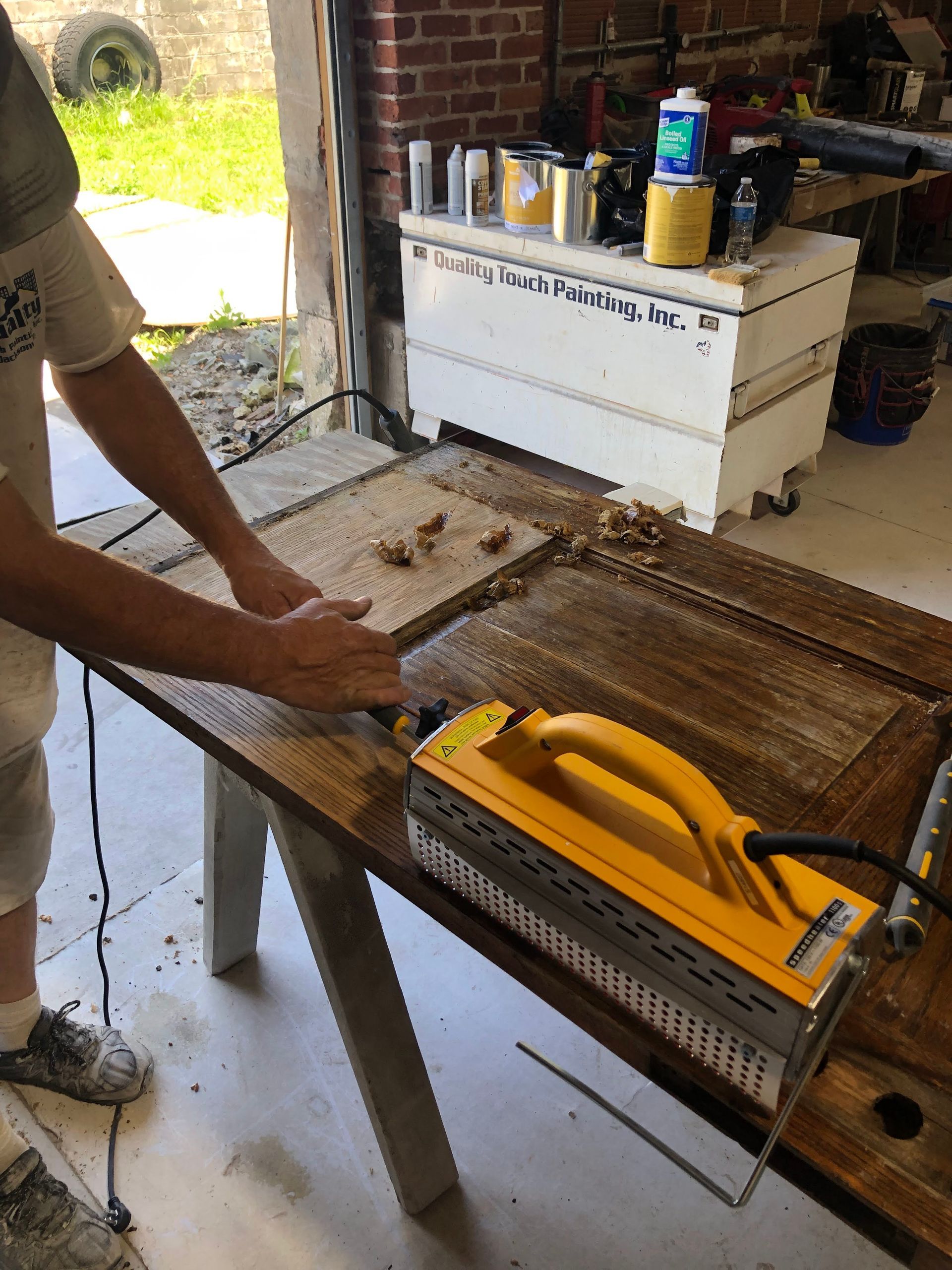 Person using a heat gun to remove finish from wooden door on a workbench outdoors.
