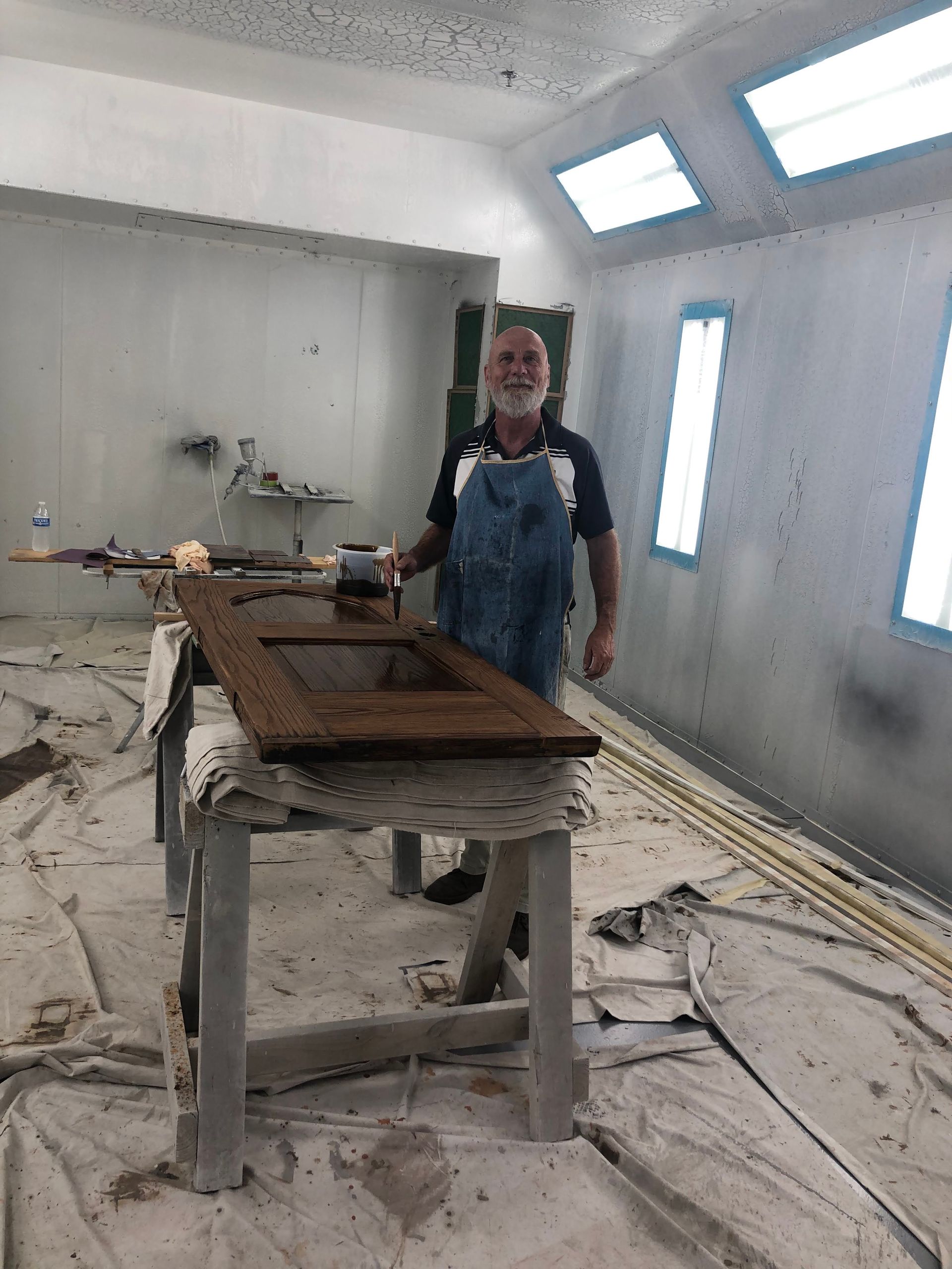 Man in apron spraying a wooden cabinet door in a paint booth.