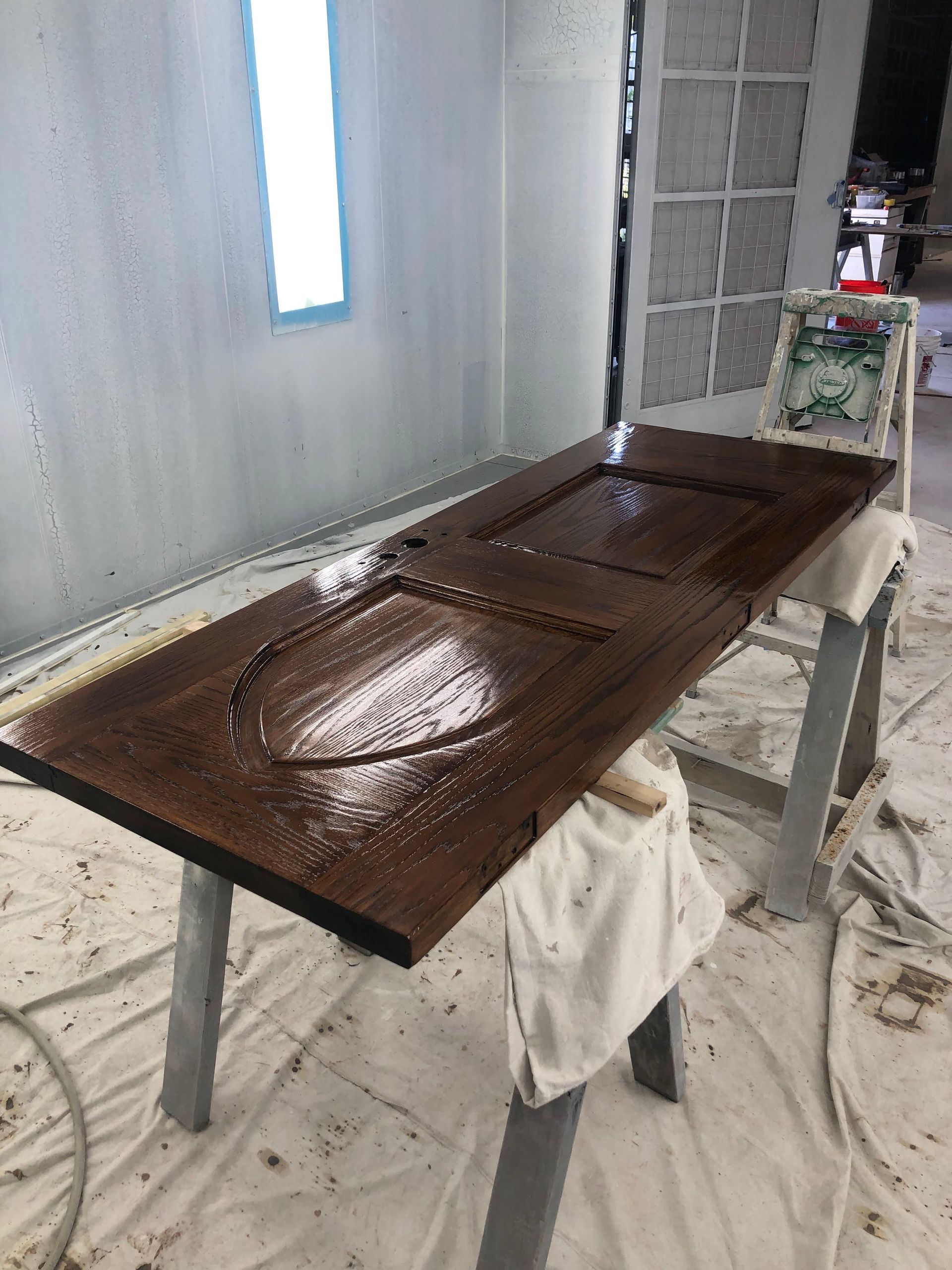 Brown door resting on saw horses, freshly coated with a glossy finish, inside a spray booth.