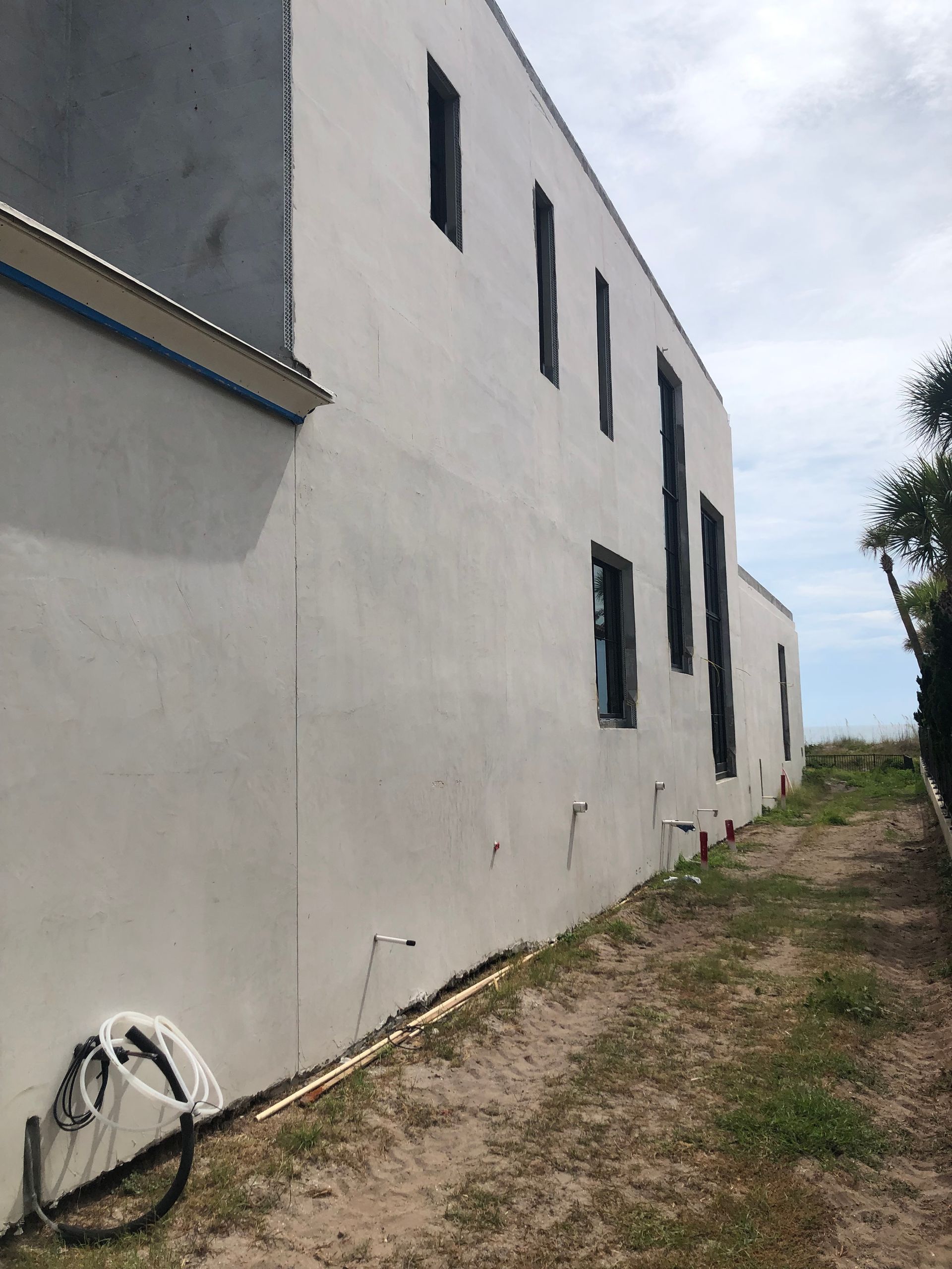 Side view of a light gray building with narrow dark windows, next to a sandy path leading to the coast.