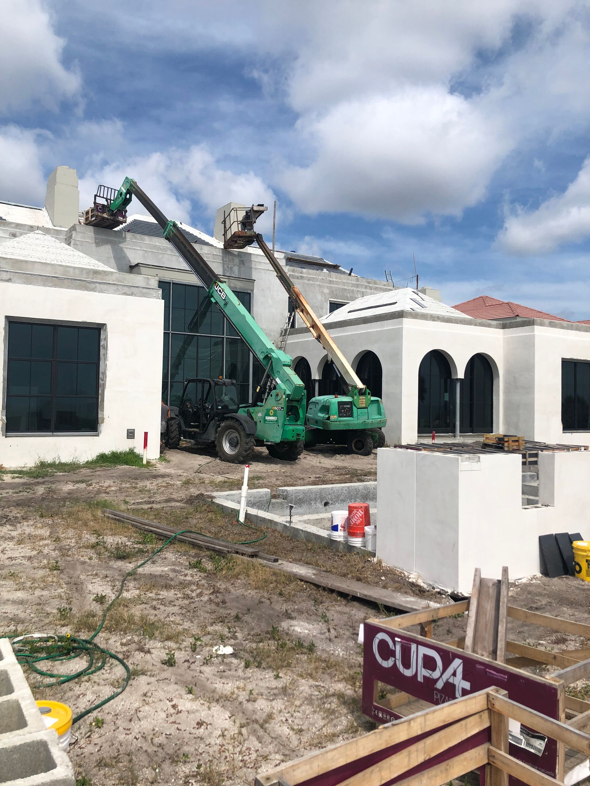 Construction site with two boom lifts near a white building with black windows, under a cloudy sky.