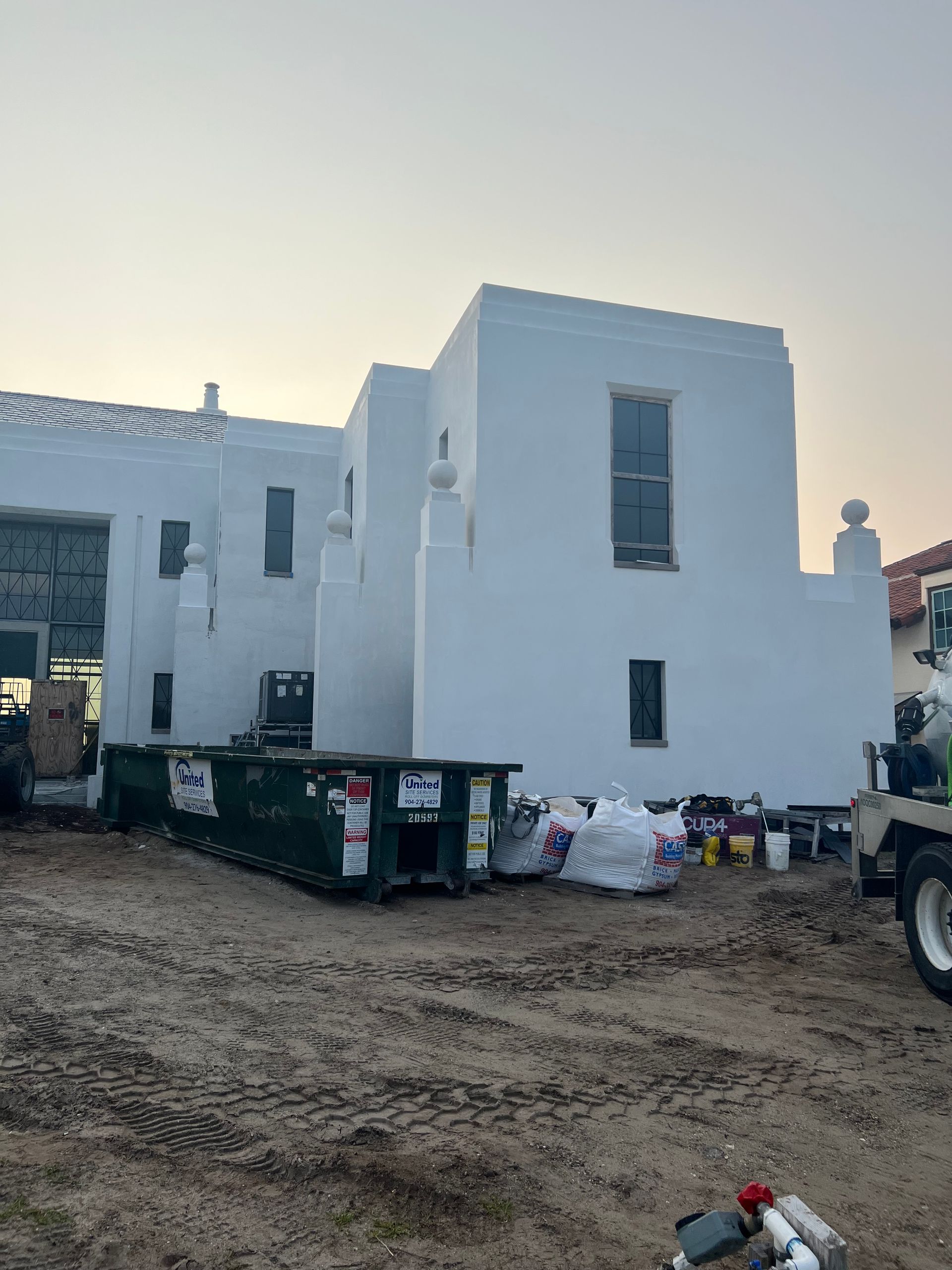 White stucco building with dumpsters, construction materials, and a dirt yard.