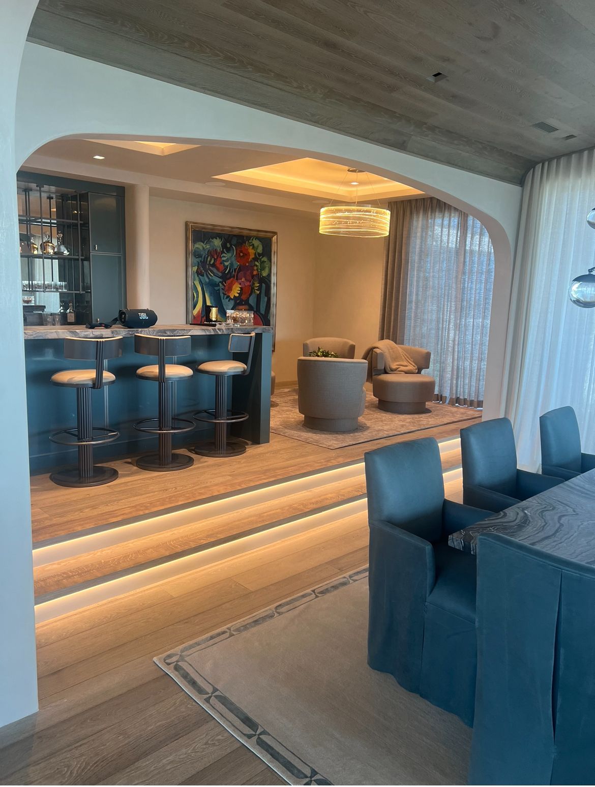 A modern living space with a bar, seating area, and dining table; teal and neutral tones.