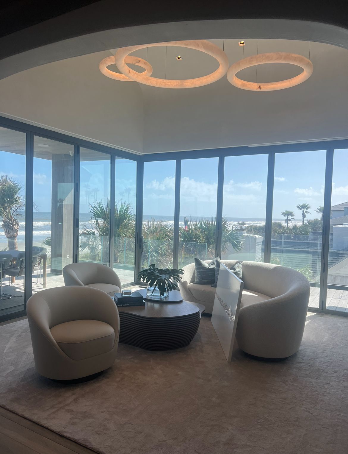 Living room with curved beige furniture, large windows overlooking the ocean, and circular ceiling lights.