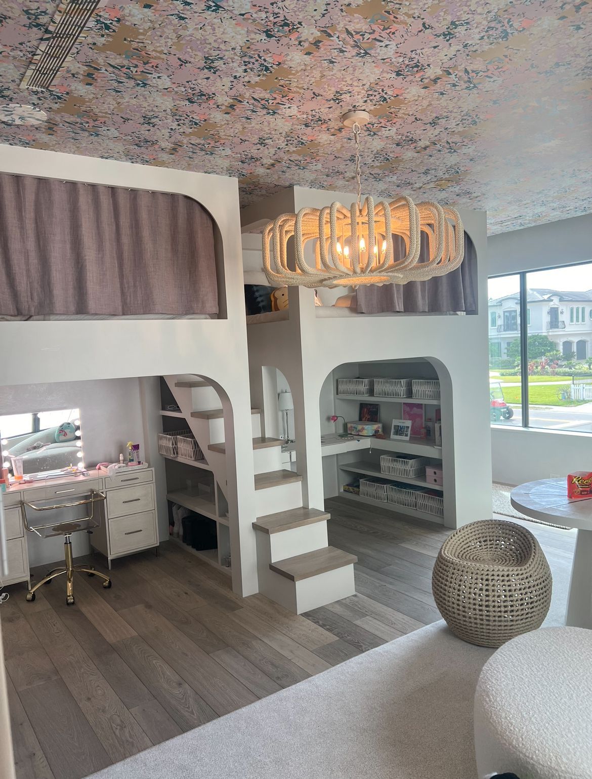 Lofted bedroom with built-in shelves and desk, light wood floors, patterned ceiling, and large chandelier.