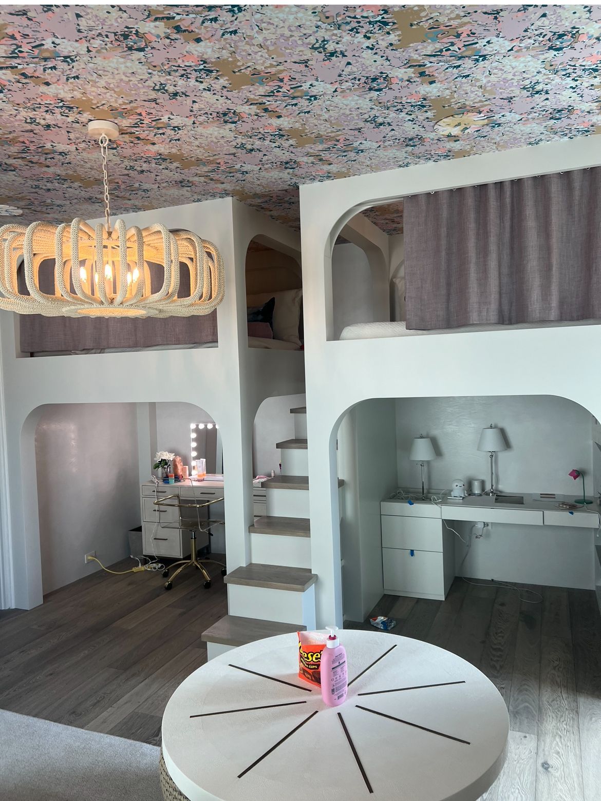 A whimsical bedroom with bunk beds, desks, and a round table, adorned with a patterned ceiling.