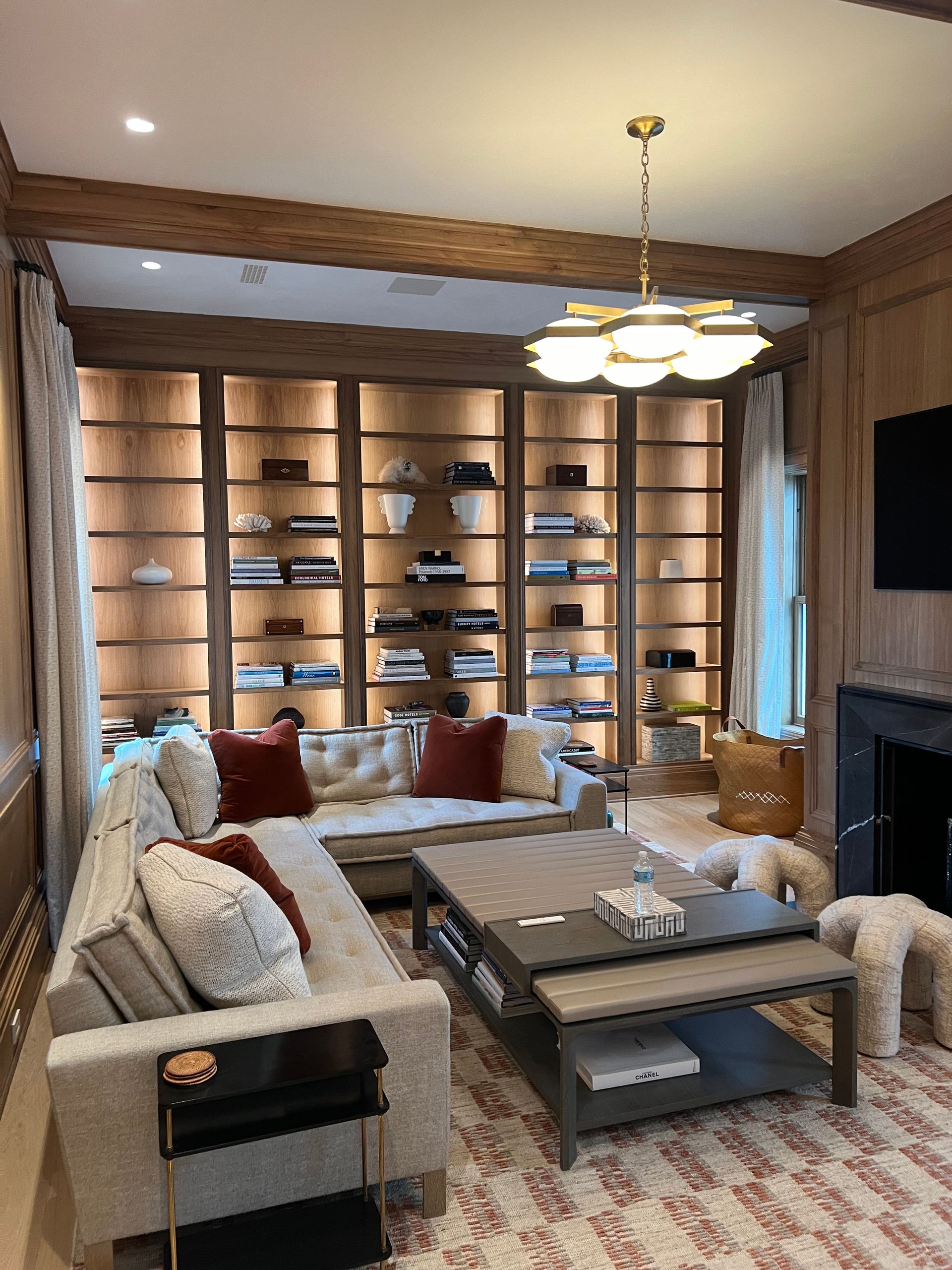 Cozy living room with built-in bookshelves, beige sofa, red pillows, wooden beams, and a coffee table on a patterned rug.