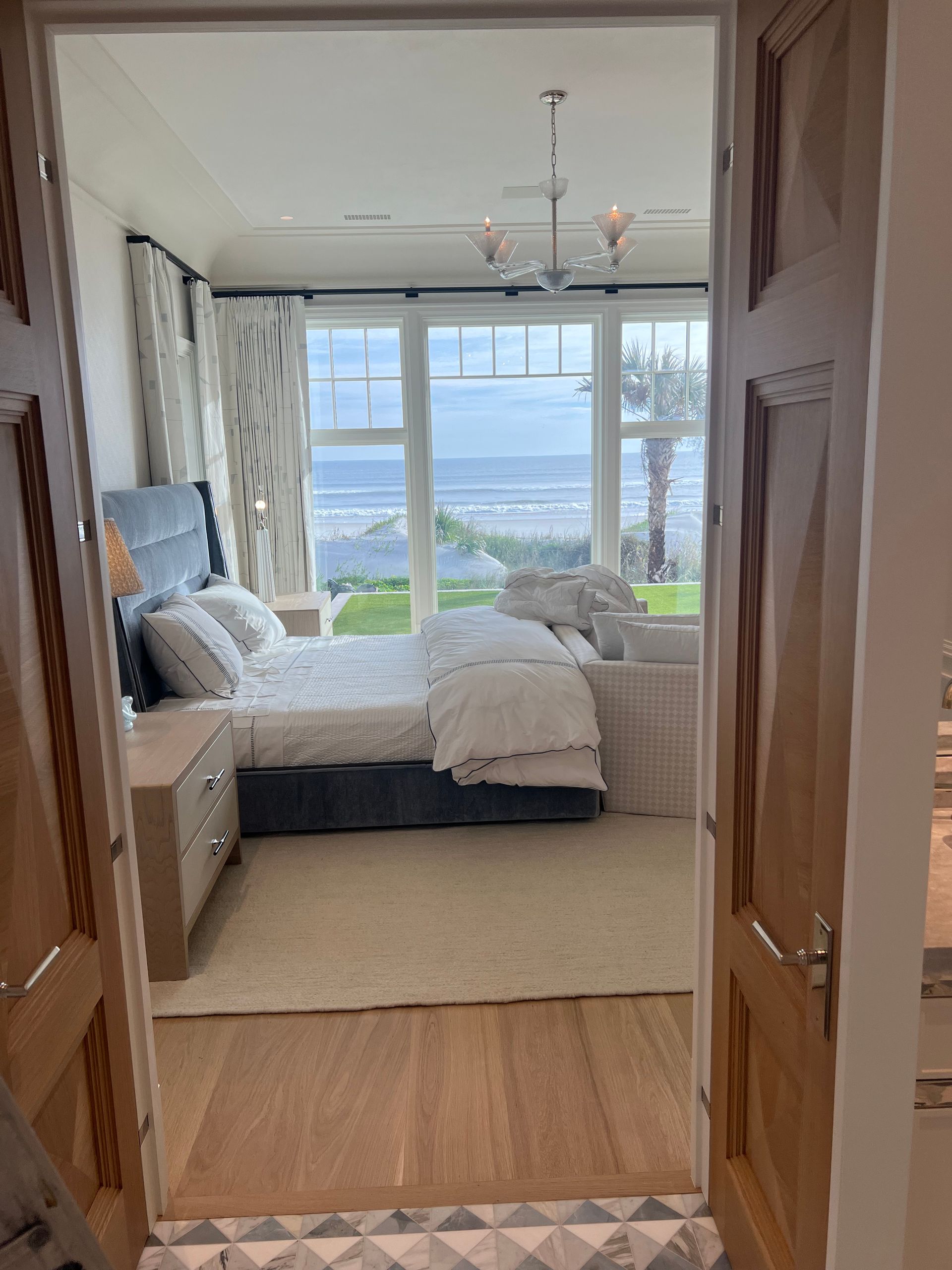 Bedroom with ocean view; bed with blue headboard, white linens; large windows, light wood floors.