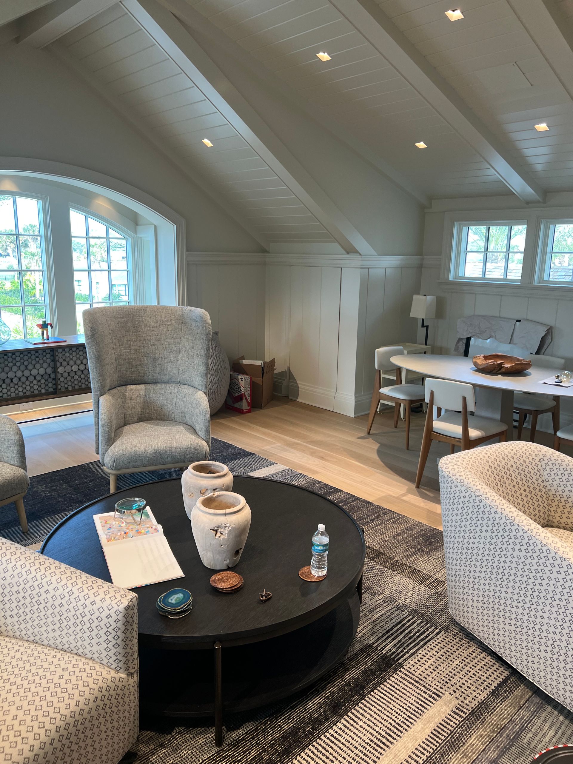 Living room with angled ceiling, patterned rug, and round coffee table. Armchairs and dining table visible.