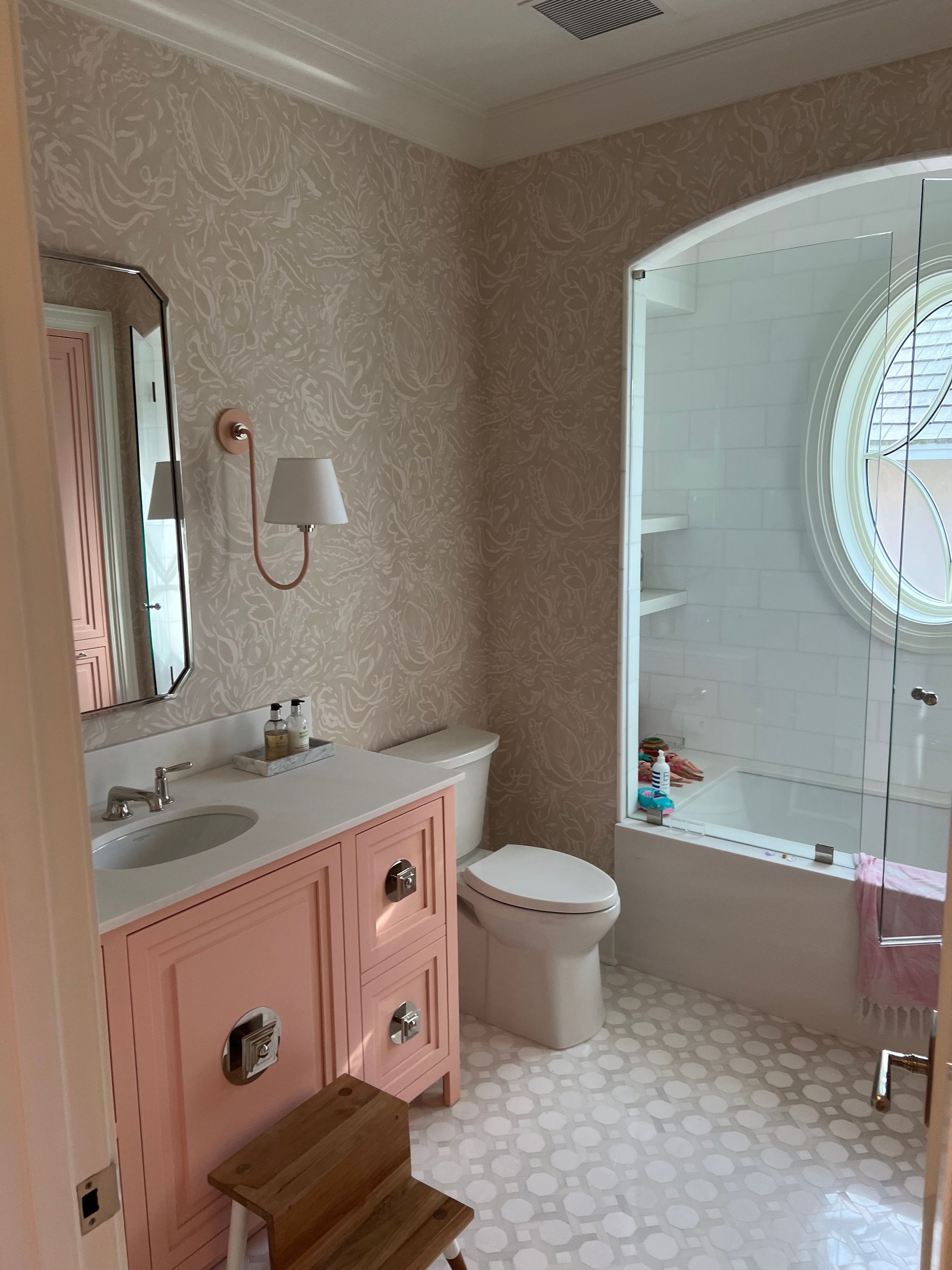 Pink bathroom with patterned wallpaper, pink vanity, oval window, and pebble tile floor.