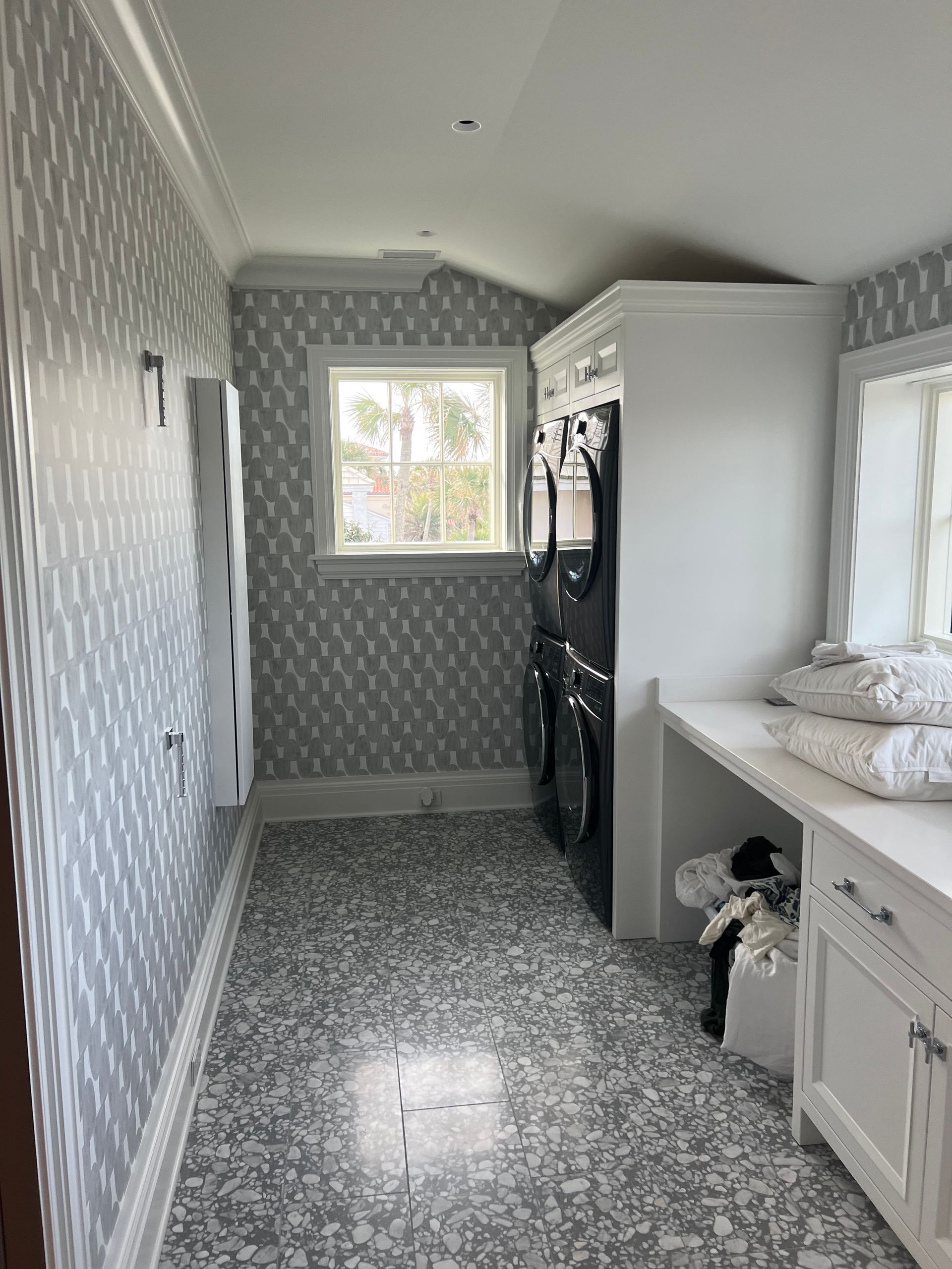 Laundry room with gray patterned wallpaper, window, white cabinets, and terrazzo flooring.