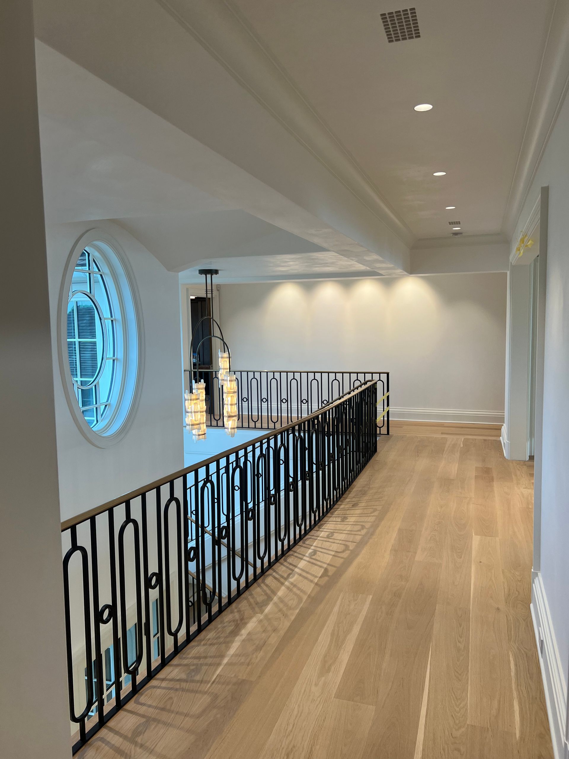 Balcony hallway with wood floors, black railing, white walls, and oval window.