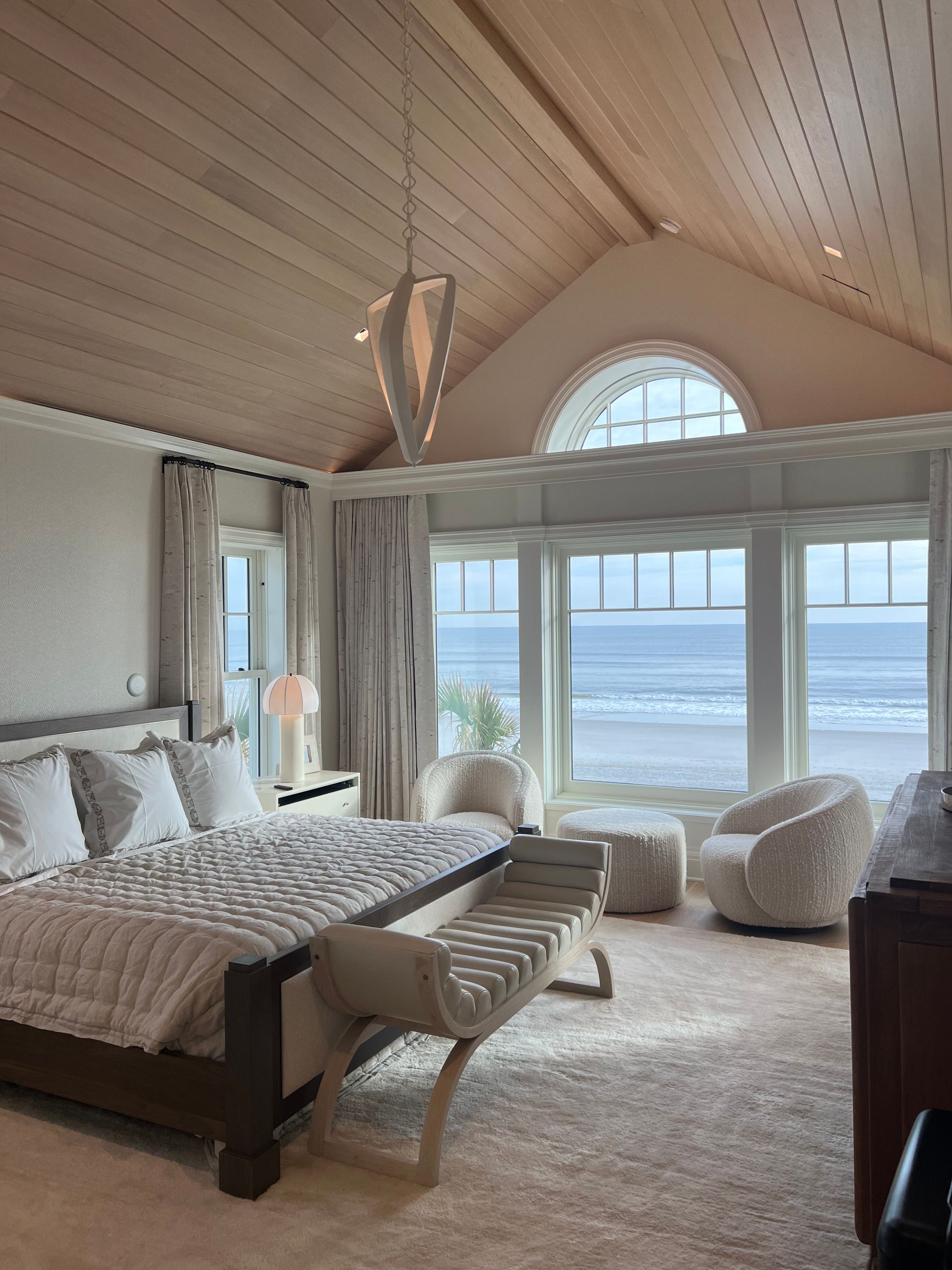 Bedroom with bed, seating, large windows overlooking beach and ocean, and light wood ceiling.