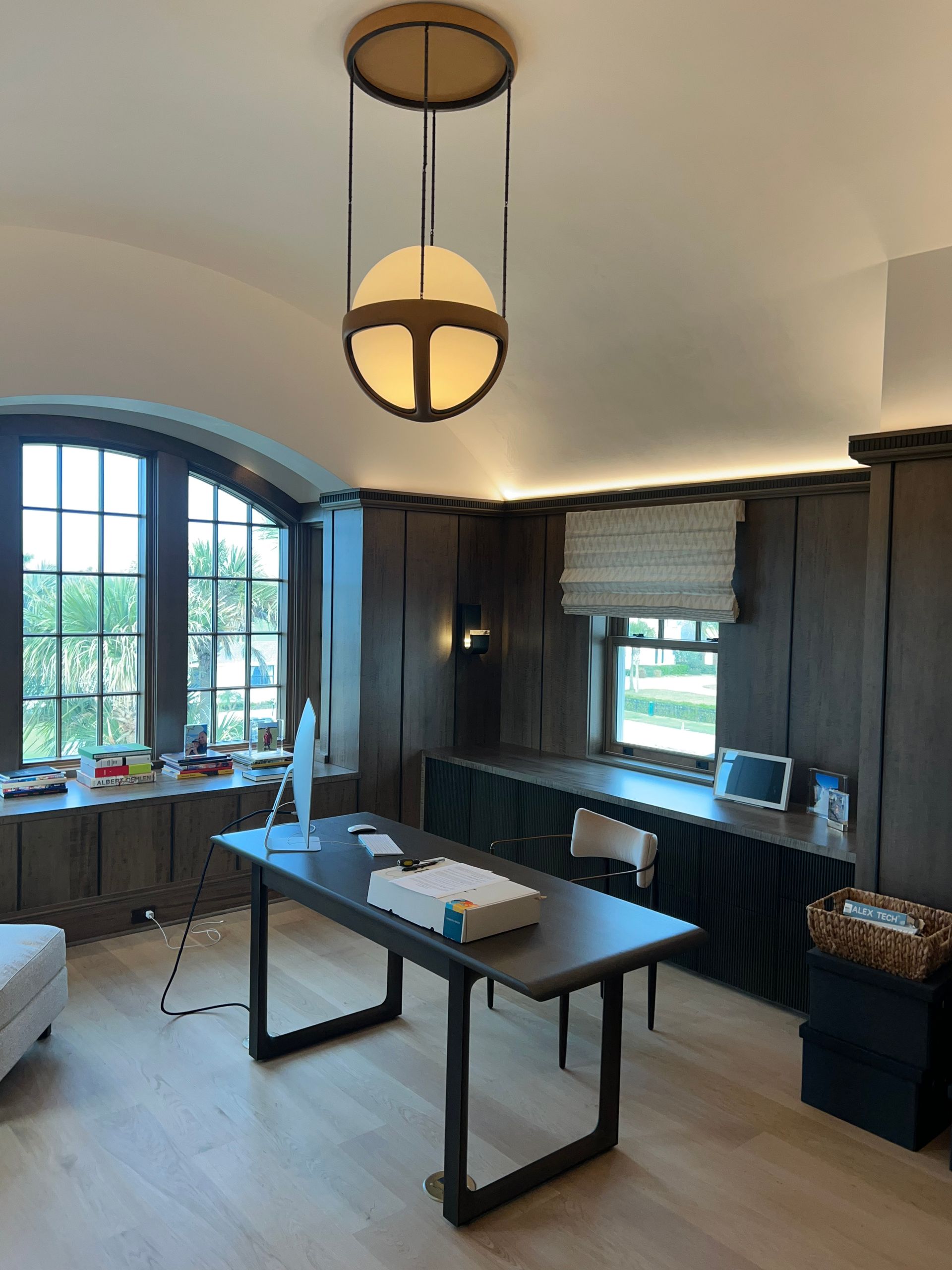 An elegant home office with a dark wood paneled wall, large desk, globe pendant light, and arched windows.