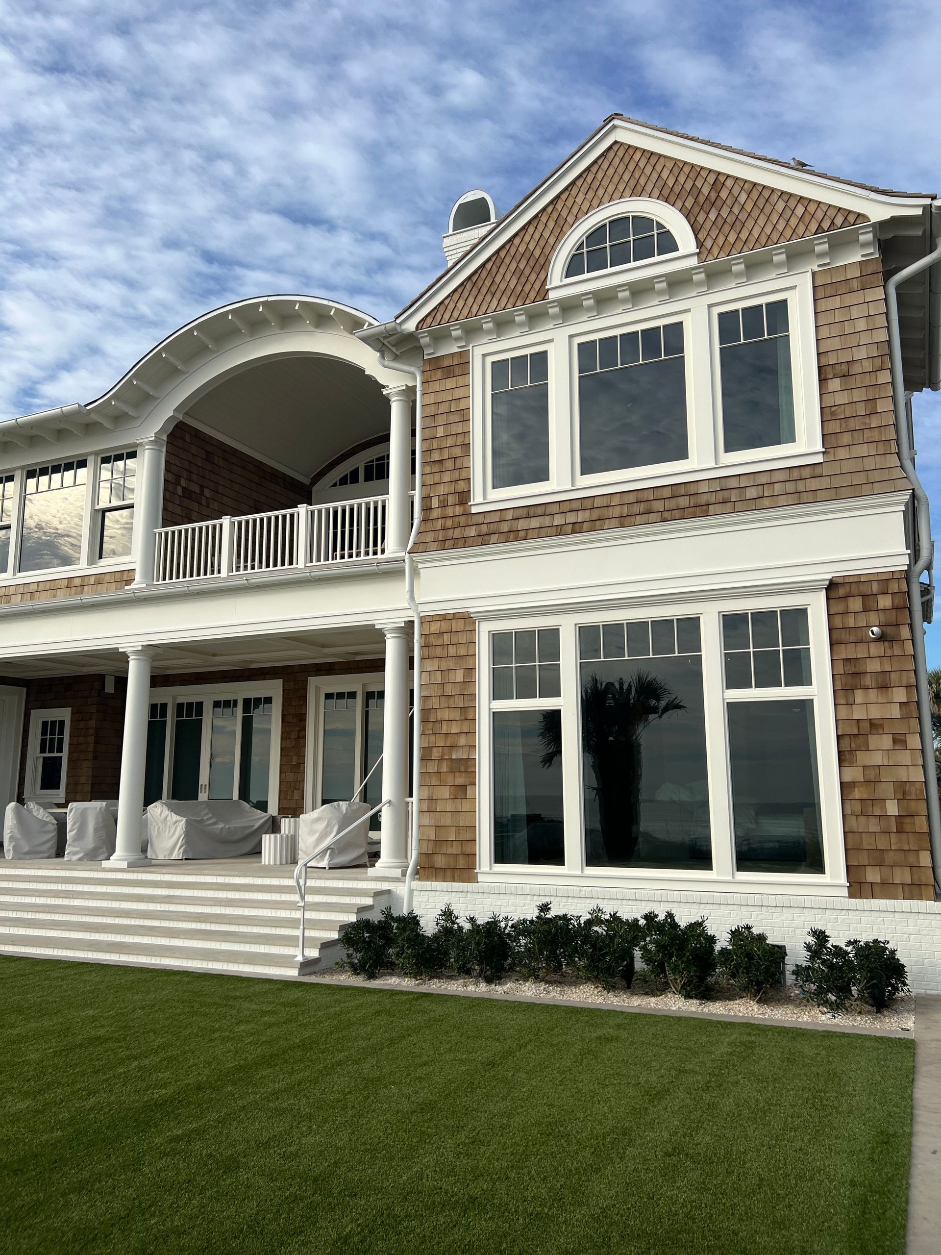 Large two-story house with brown shingle siding, white trim, and columns, next to green lawn under a cloudy sky.
