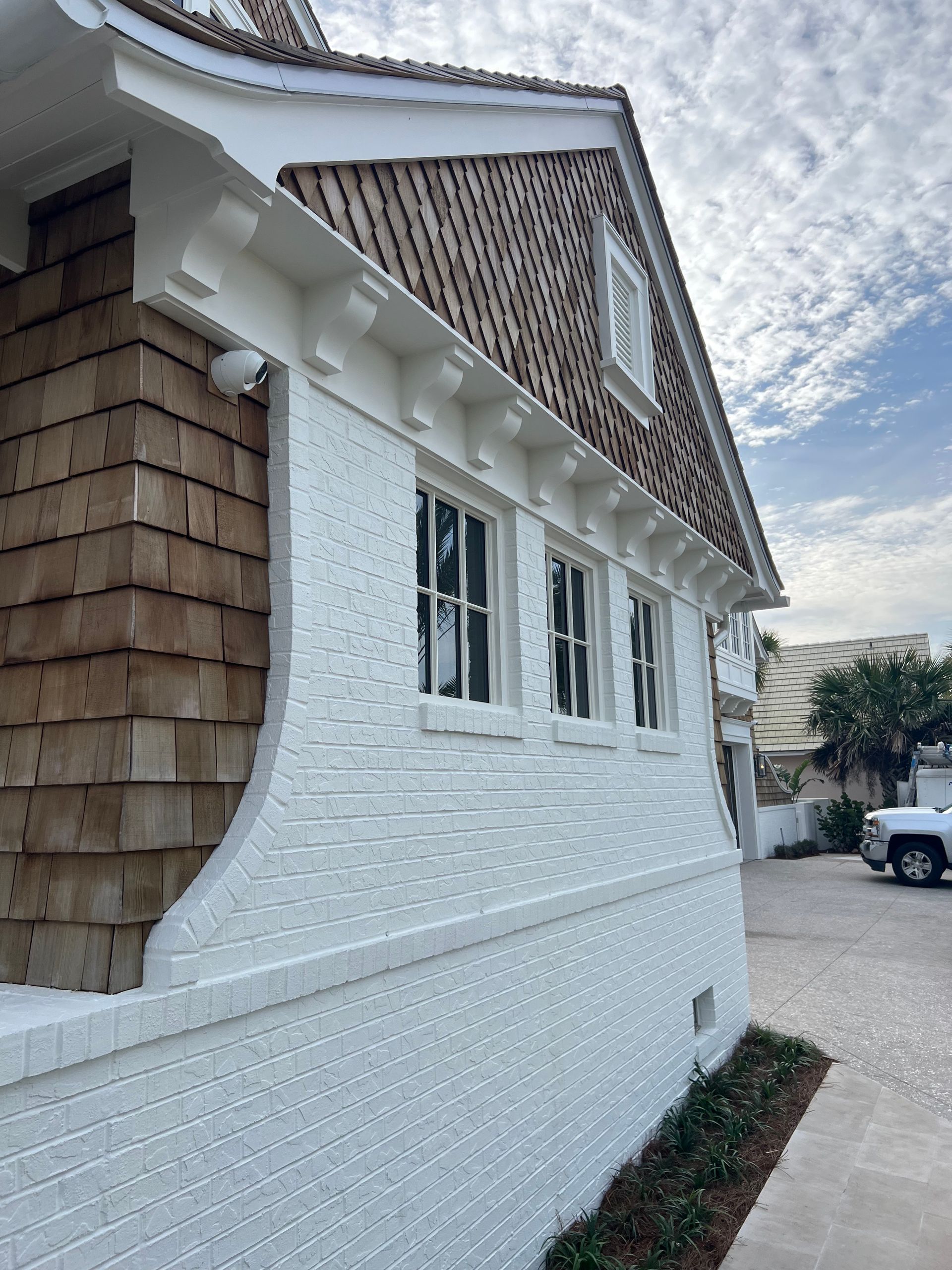 White brick building with brown shingle siding and three windows, beneath a cloudy sky.